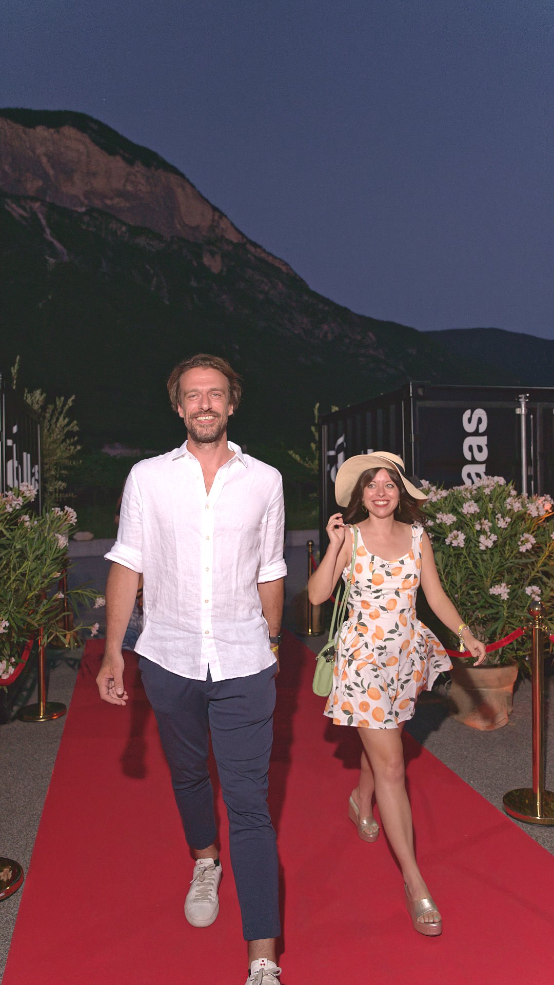 Couple in summer outfits on a red carpet below the mountains at an evening event