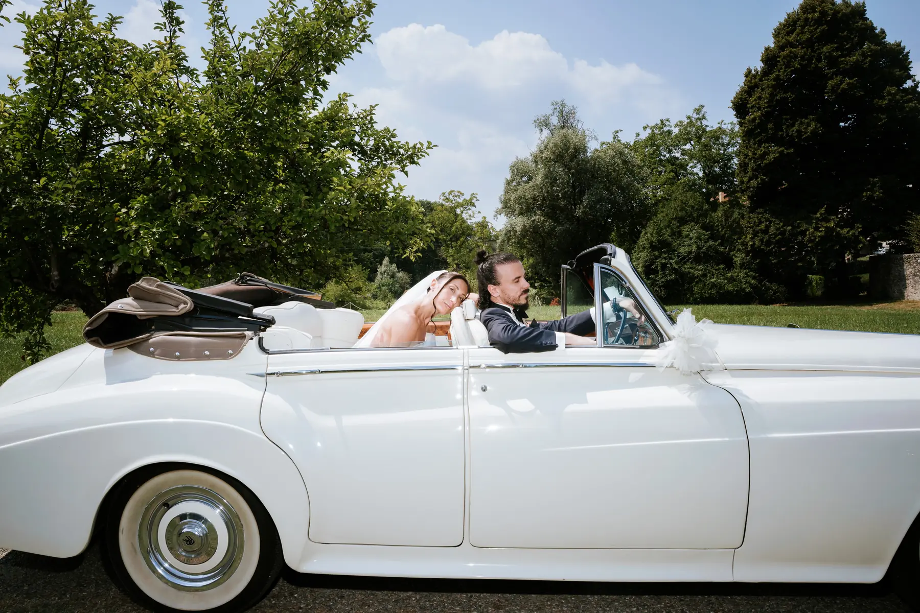 Bride and groom riding in a white vintage Rolls-Royce through the villa park