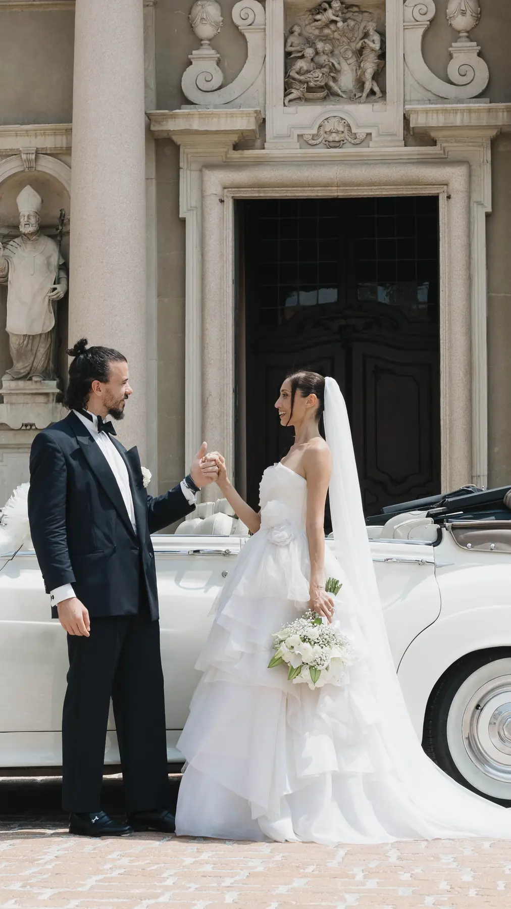 Bride and groom facing each other beside the vintage car at the villa entrance
