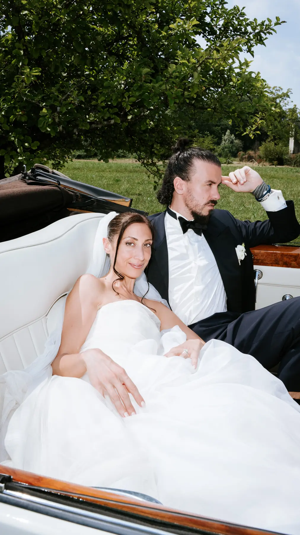 Newlyweds seated in the vintage convertible under the trees, wedding celebration