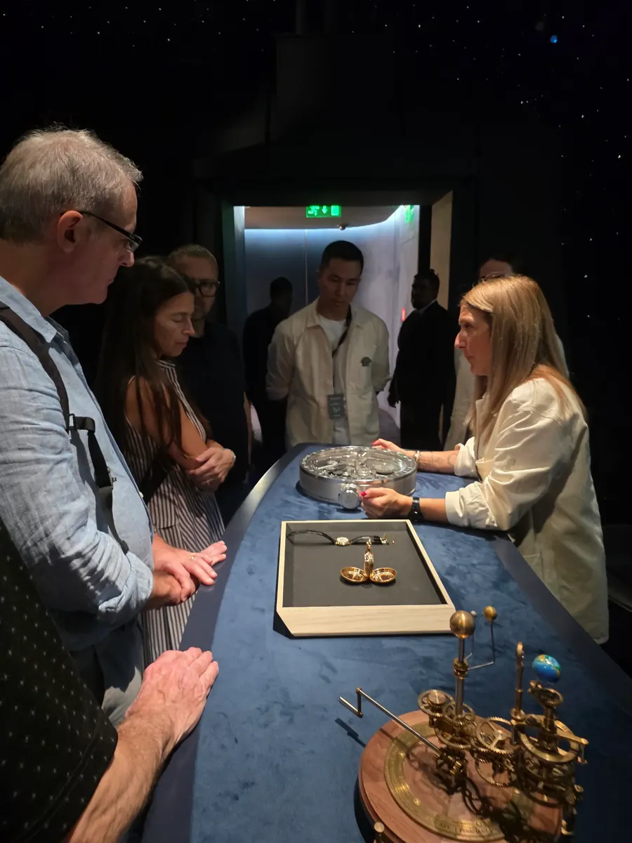 Guests around a blue velvet table listening to a watchmaking presentation, an orrery in the foreground
