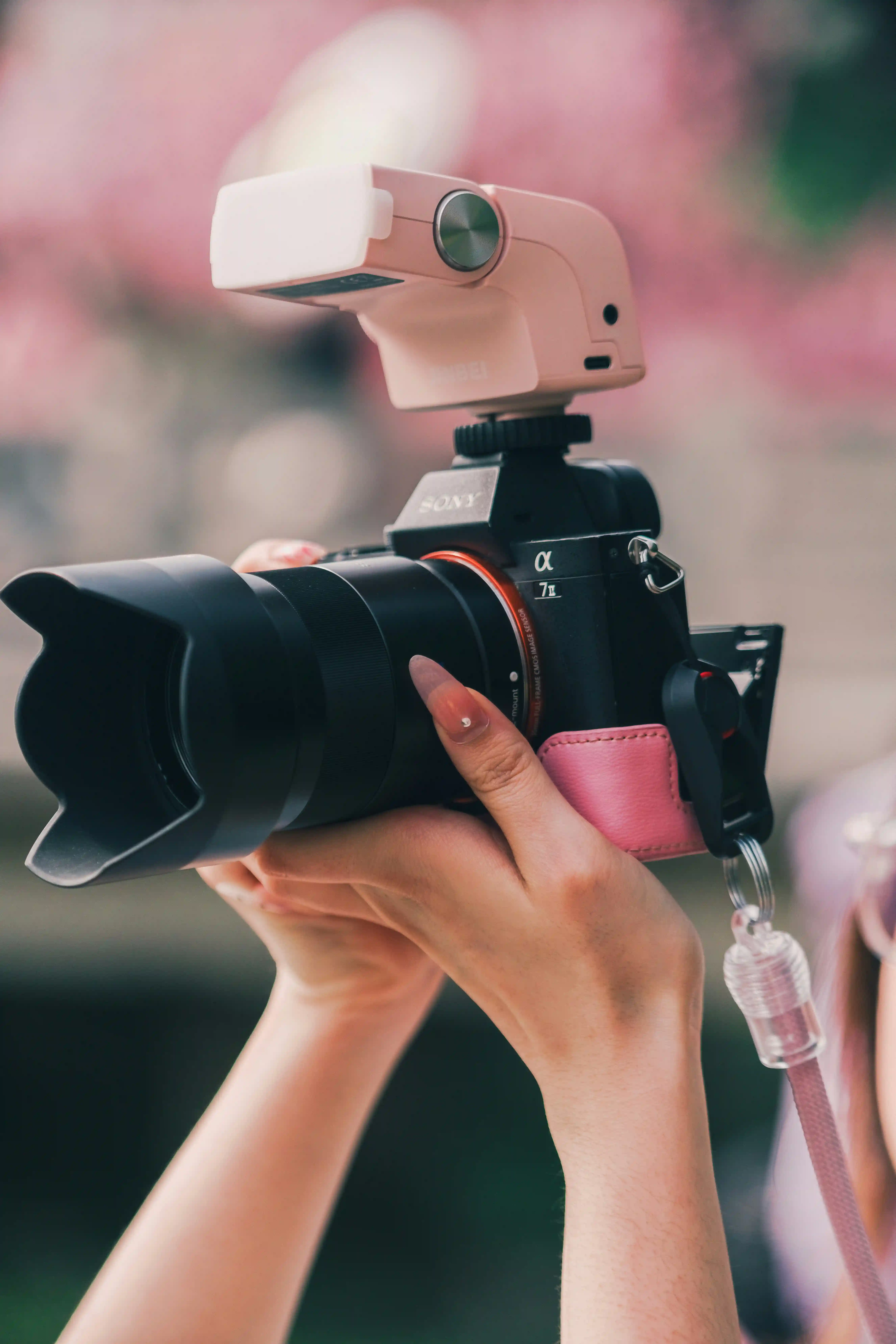 Creative photographer talent holding Sony camera with pink strap in cherry blossom setting for Coverpage Dubai showcase.