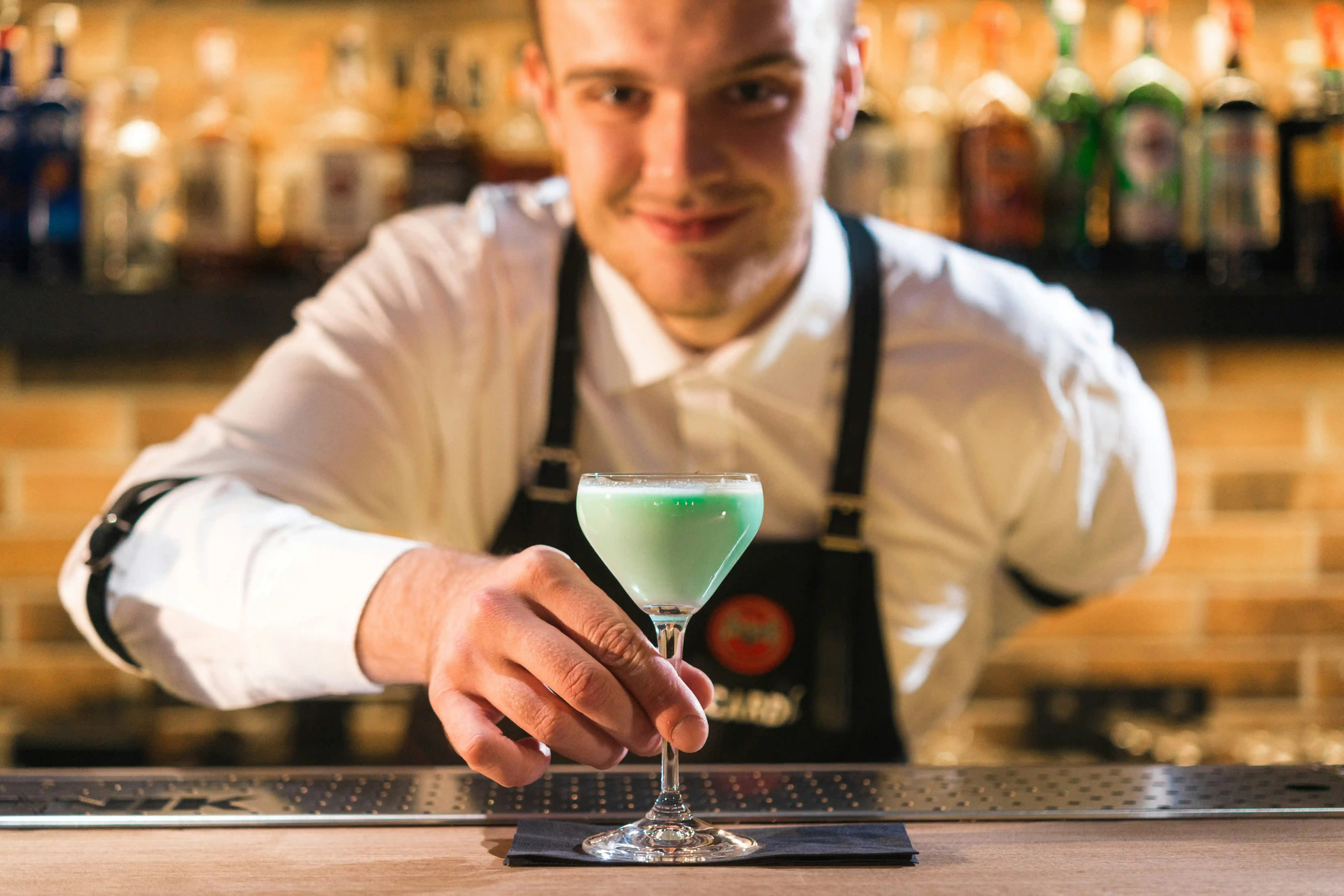 Smiling bartender talent holding green cocktail glass at bar for Coverpage Dubai luxury entertainment agency.