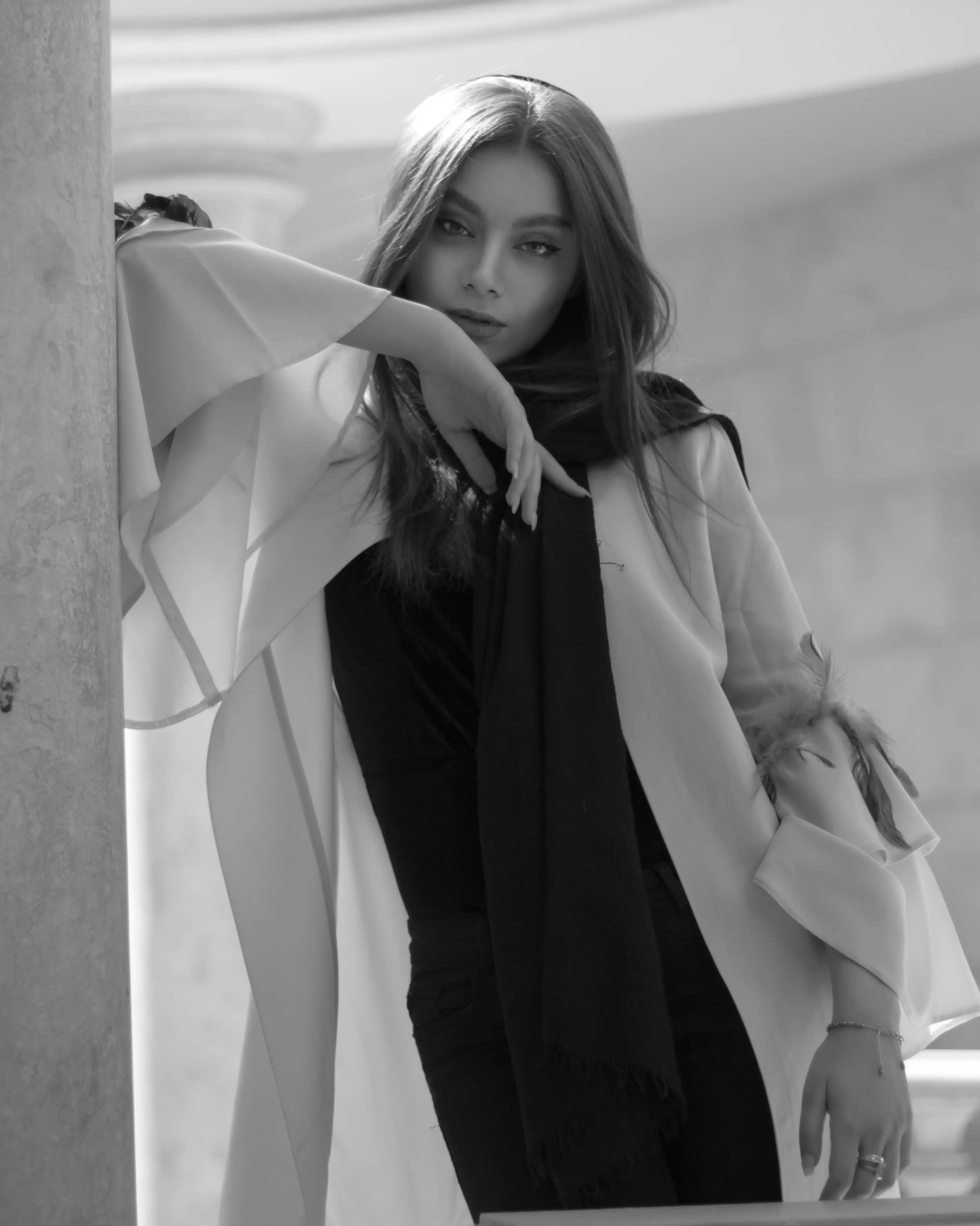 Elegant model talent leaning on pillar in white coat and black scarf for Coverpage Dubai luxury fashion showcase.