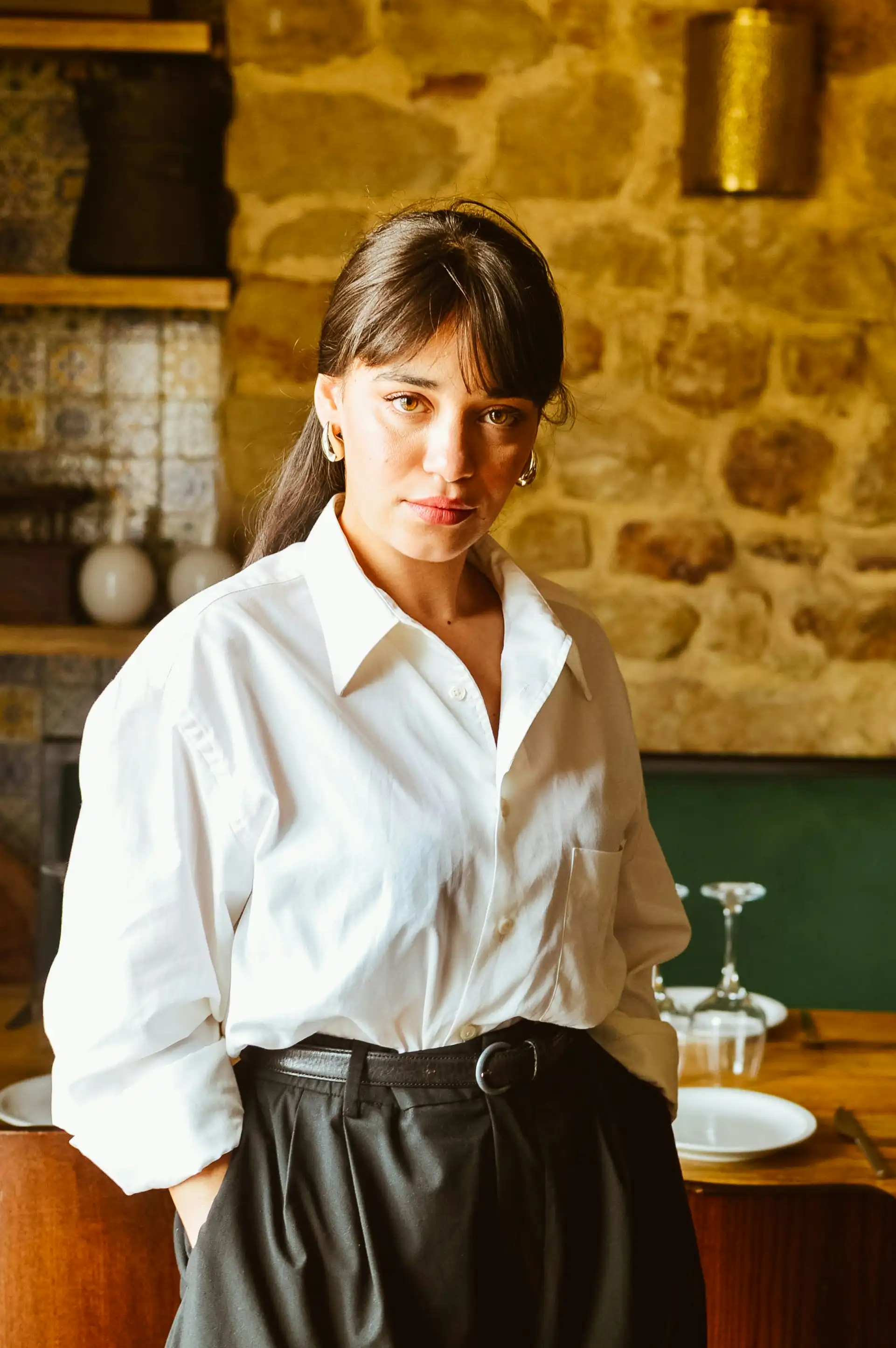Cover Page stylish waitress in white button-up shirt and black pants, hands in pockets, standing in rustic restaurant with stone wall and table setup.