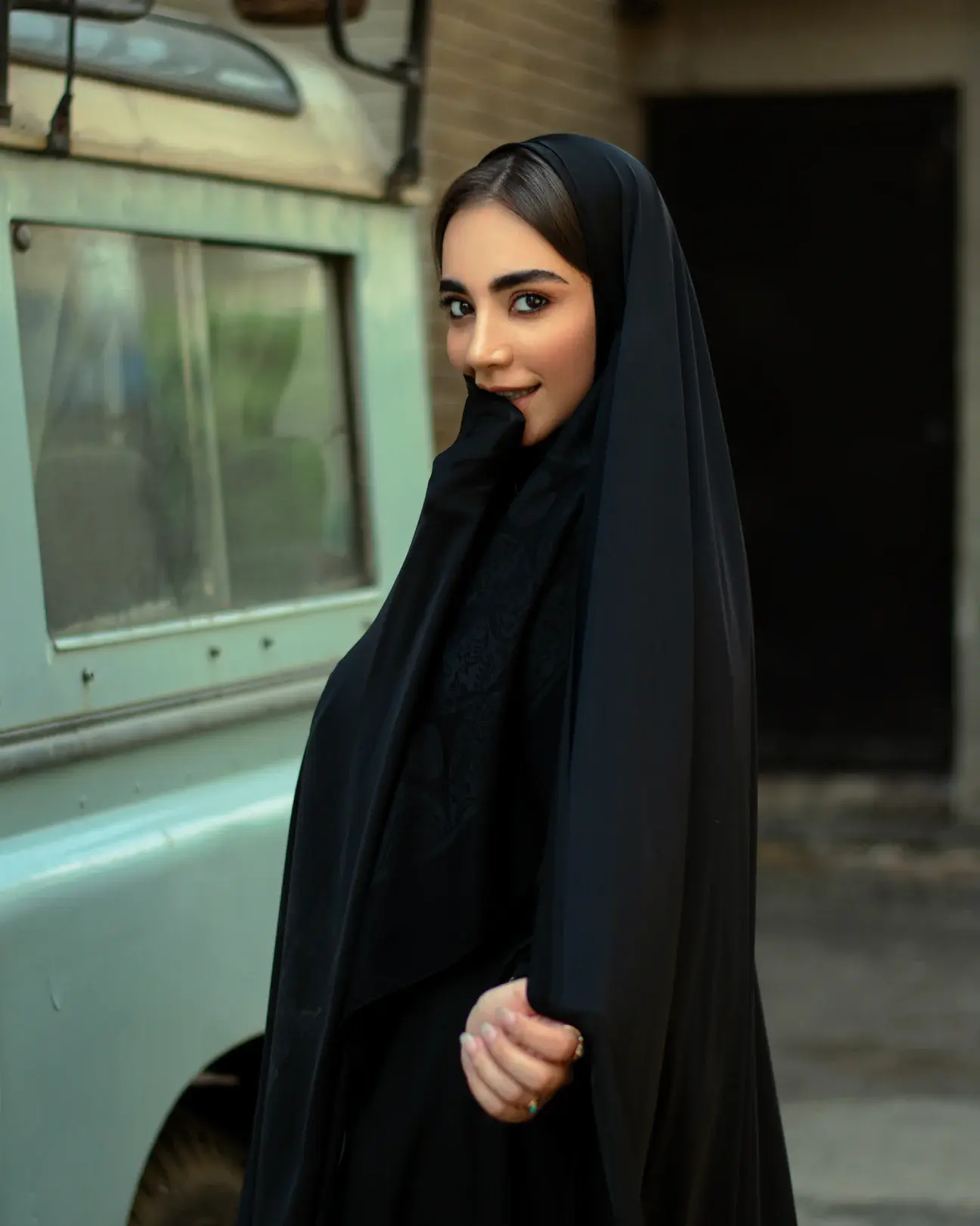 Cover Page model in black abaya and hijab, smiling with hand near mouth, standing beside vintage green vehicle in outdoor setting.