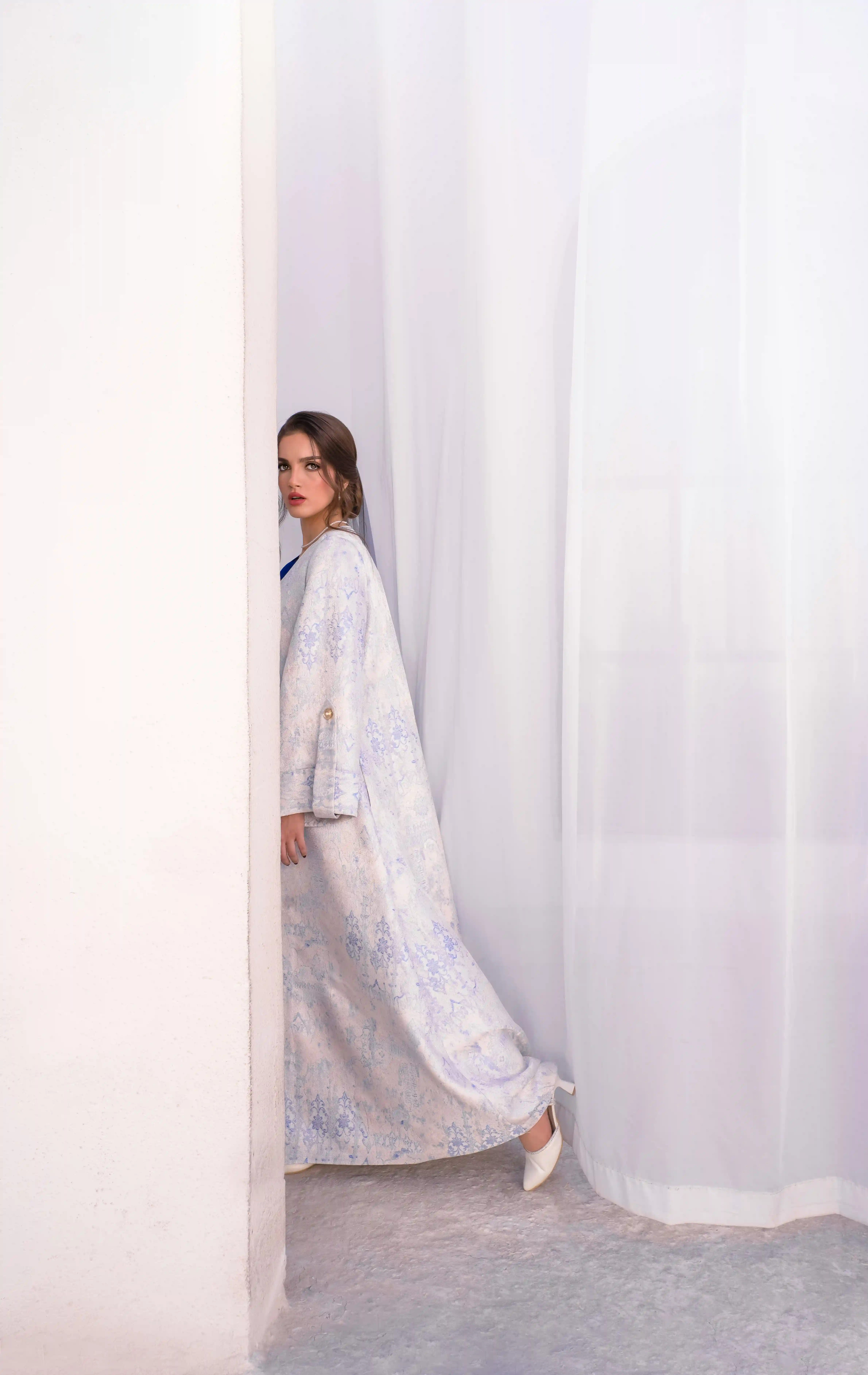 Cover Page elegant model in light blue patterned abaya and white heels, peeking around white curtain corner in bright interior.