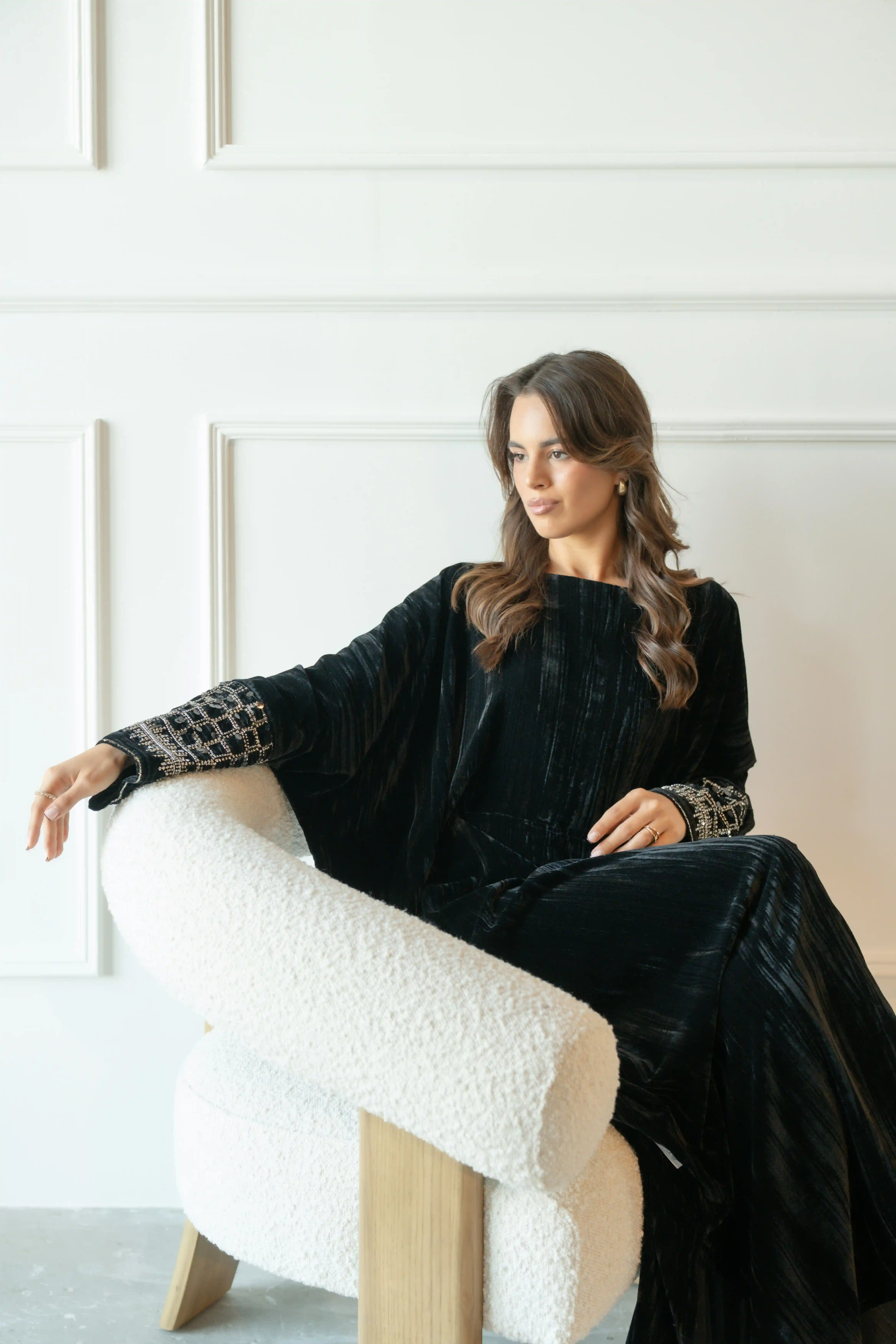 Cover Page model in black velvet abaya with embellished sleeves, reclining on modern white armchair in sophisticated white room setting.