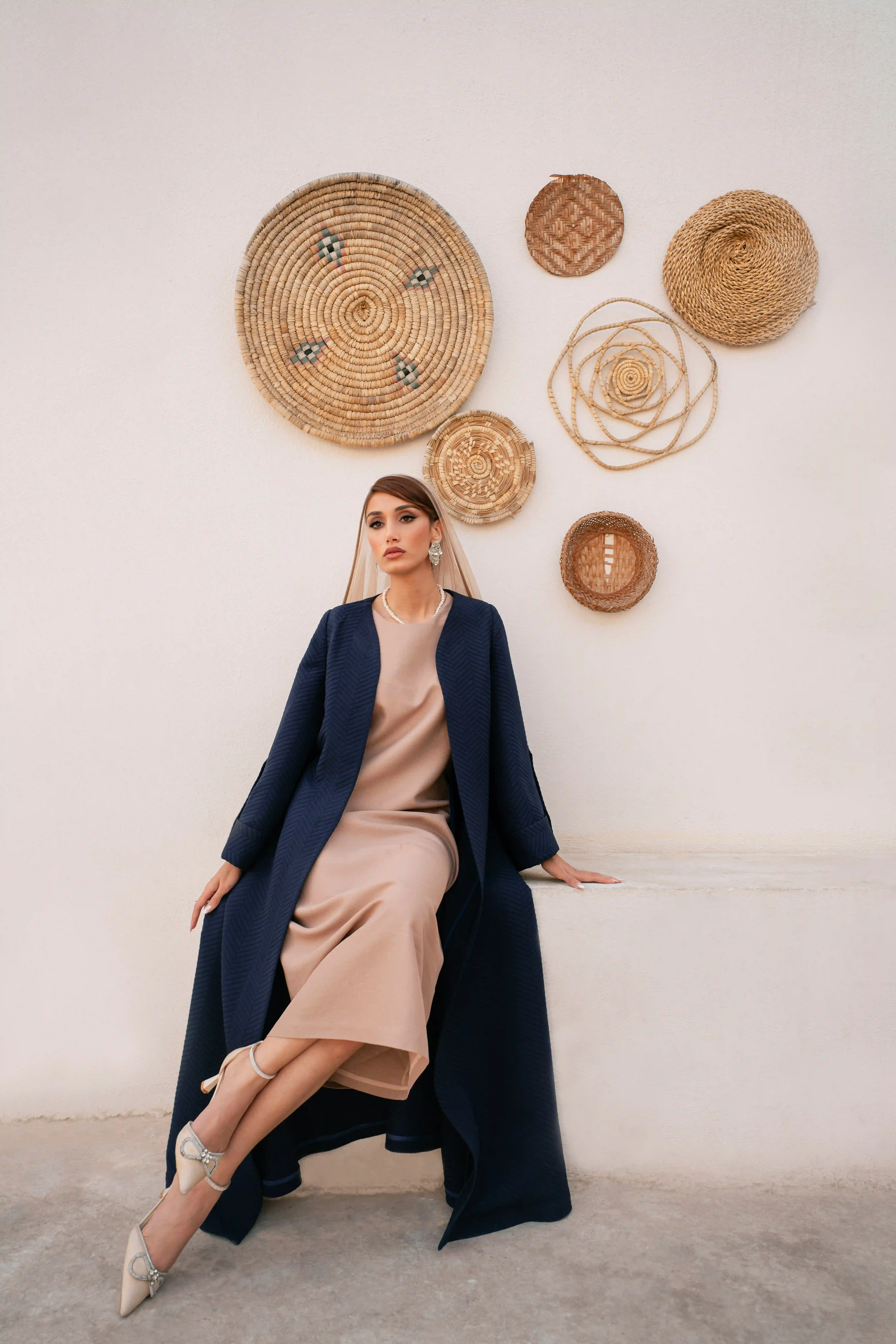 Cover Page model in beige dress and navy abaya with hijab, pearl necklace, sitting elegantly on white bench against wall with woven baskets.