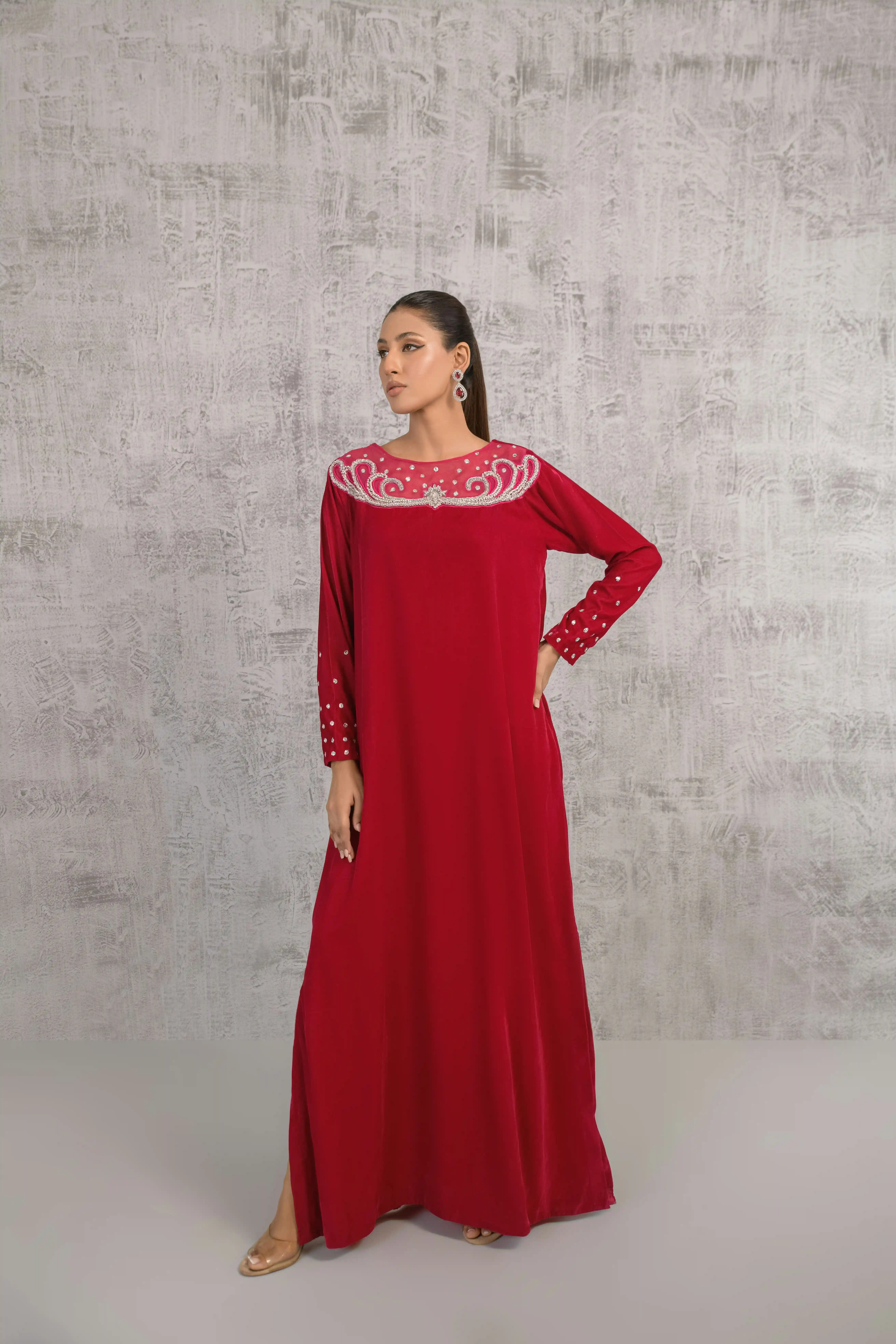Cover Page model in deep red embellished abaya, standing with hand on hip in full-body pose against textured gray wall.
