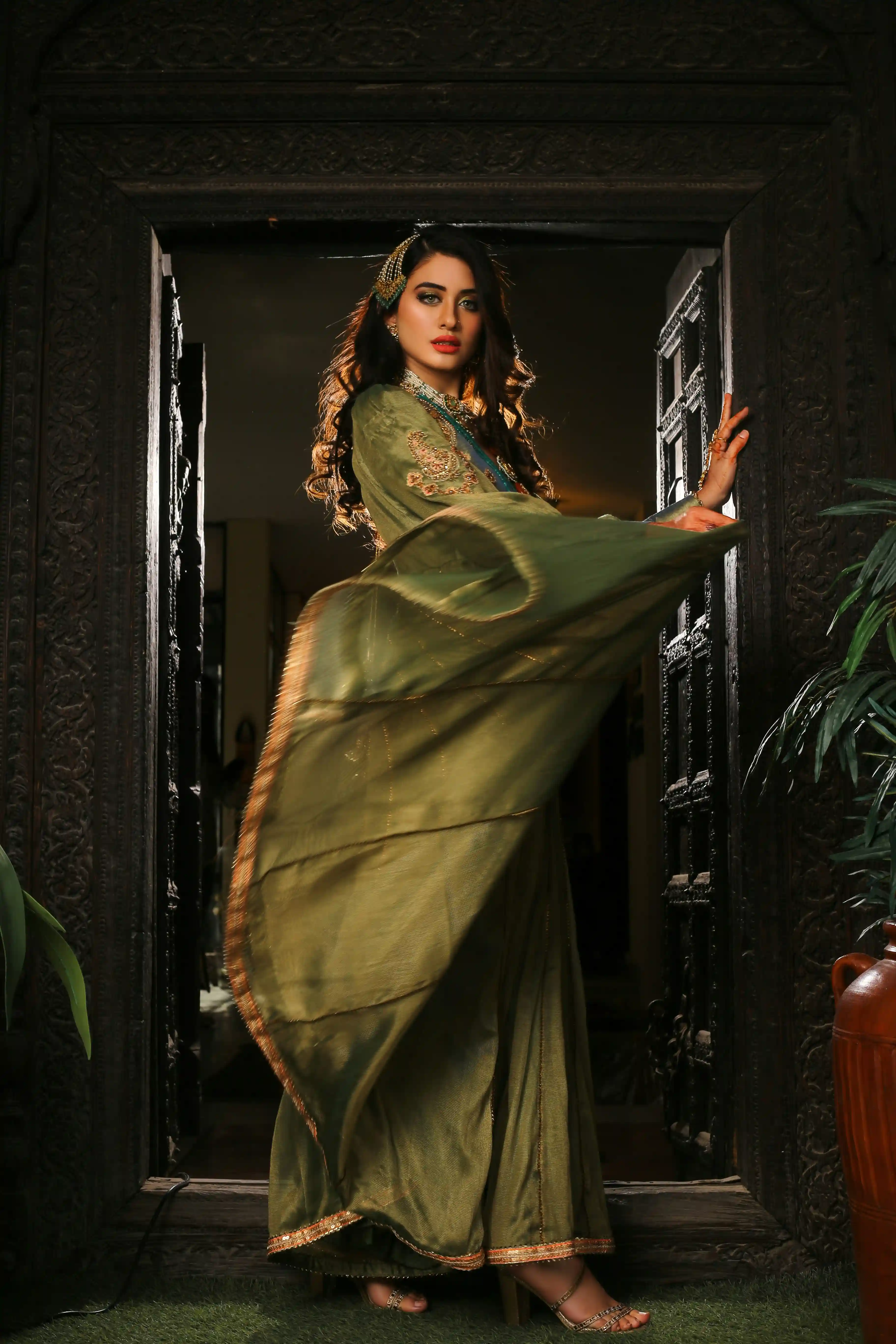 Cover Page model in green embroidered abaya and dupatta with jewelry, standing in ornate wooden doorway with flowing fabric.