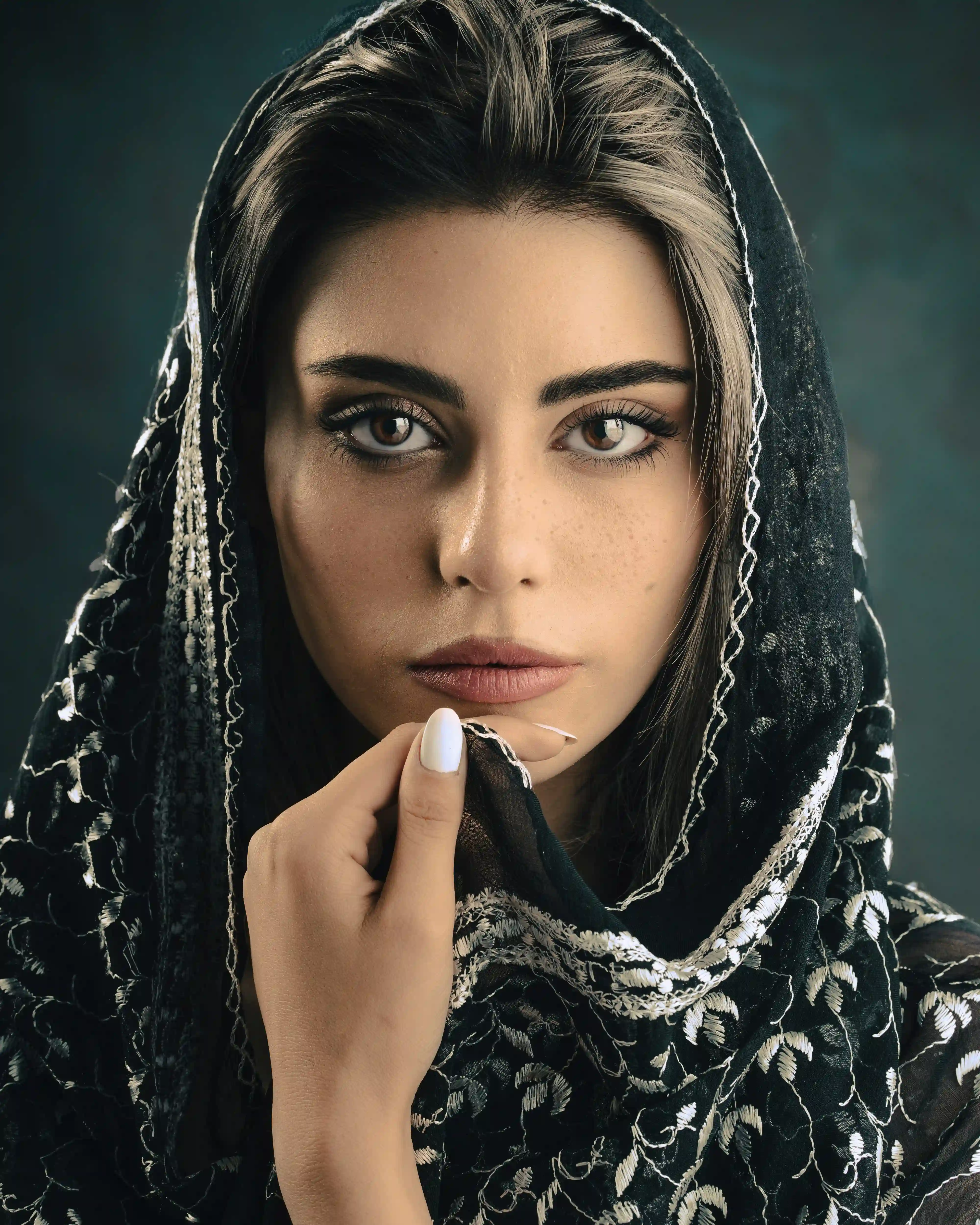 Cover Page model in black embroidered hijab and outfit, adjusting veil with hand near chin, close-up on teal background.