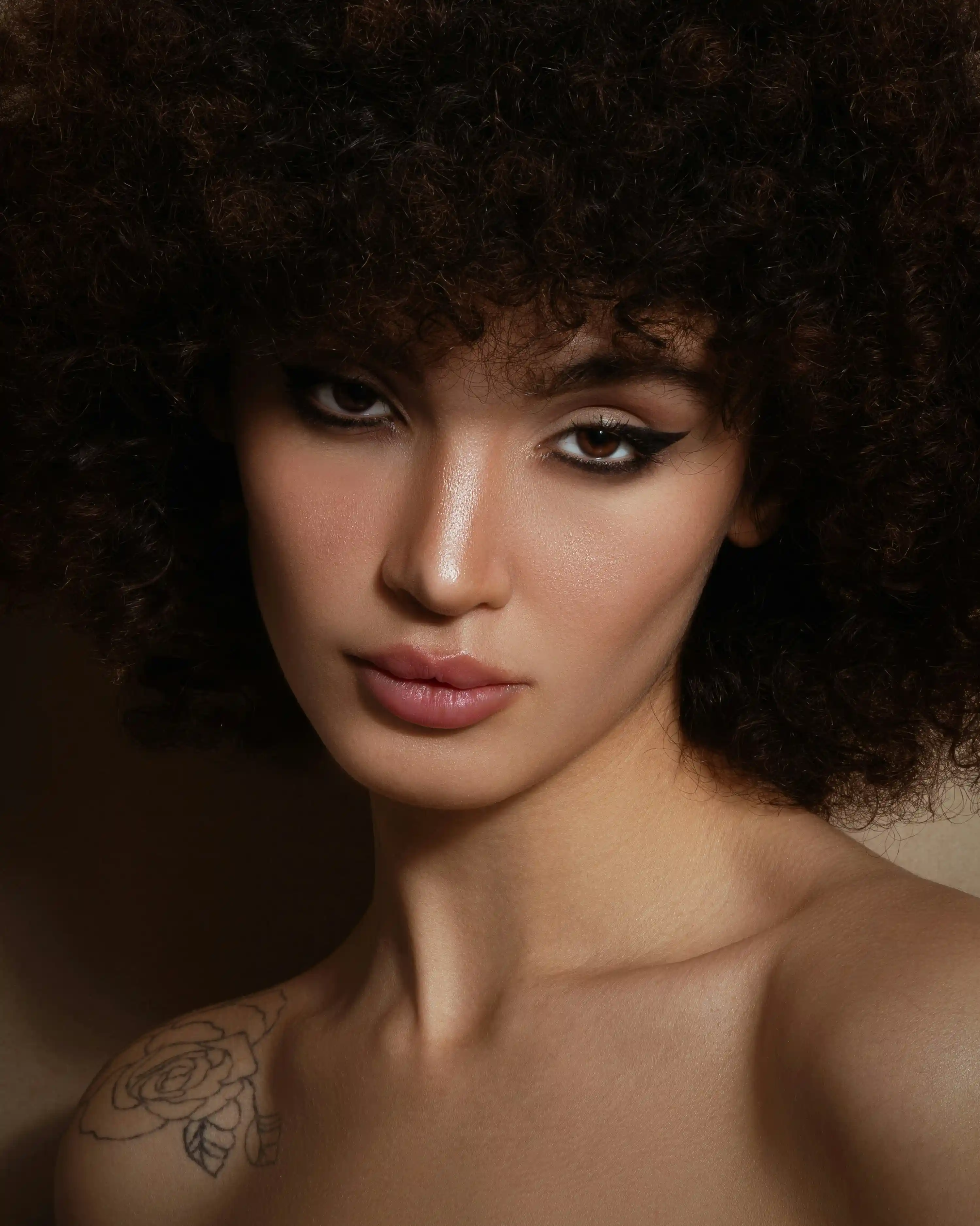 Cover Page model with curly afro hair and rose tattoo on shoulder, natural makeup with winged eyeliner, on warm background.