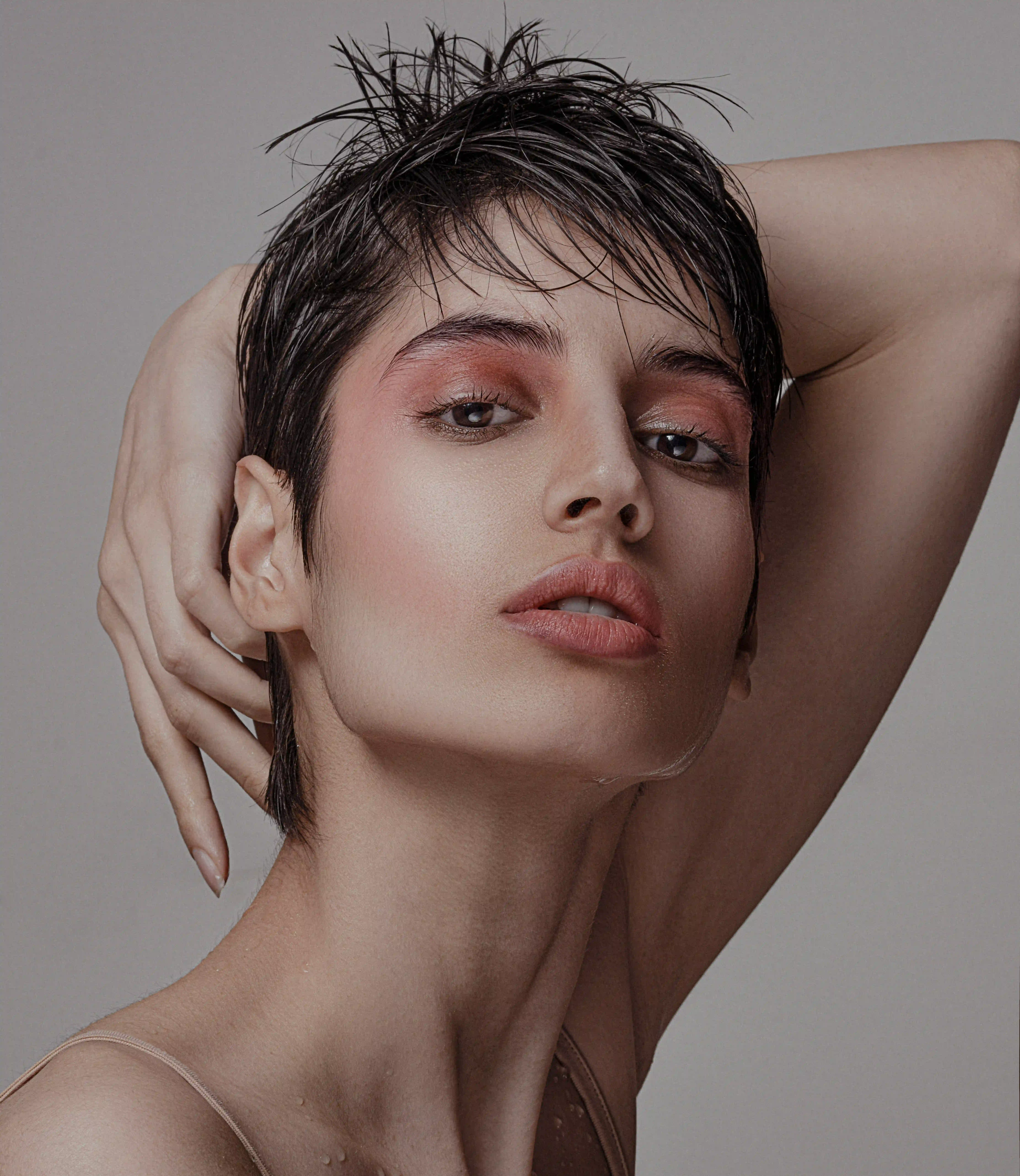Cover Page model with wet short hair, pink eyeshadow, hand on head in dramatic pose on gray background.
