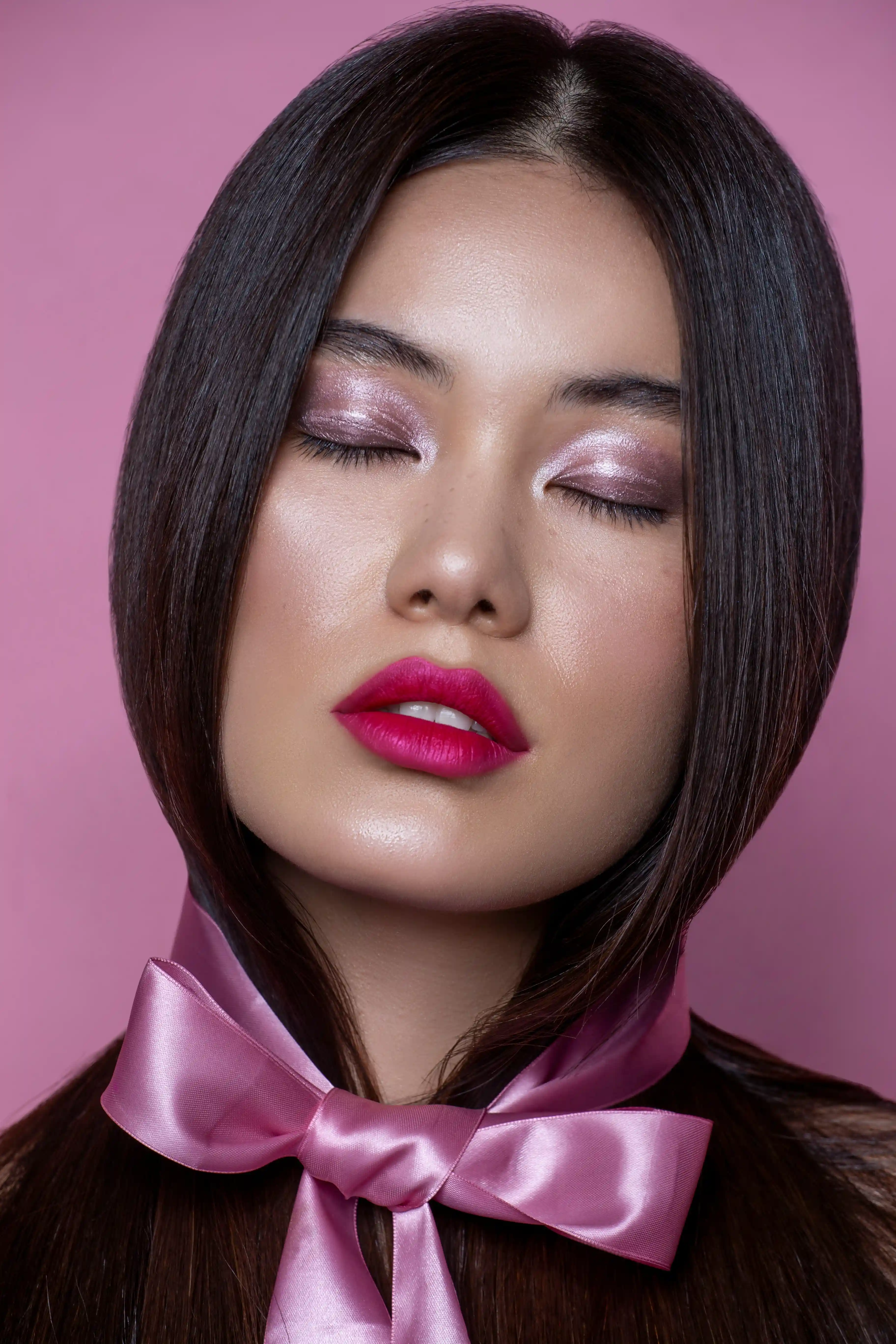 Cover Page model with straight hair and pink ribbon bow tie, closed eyes with purple eyeshadow and red lips, on pink background.