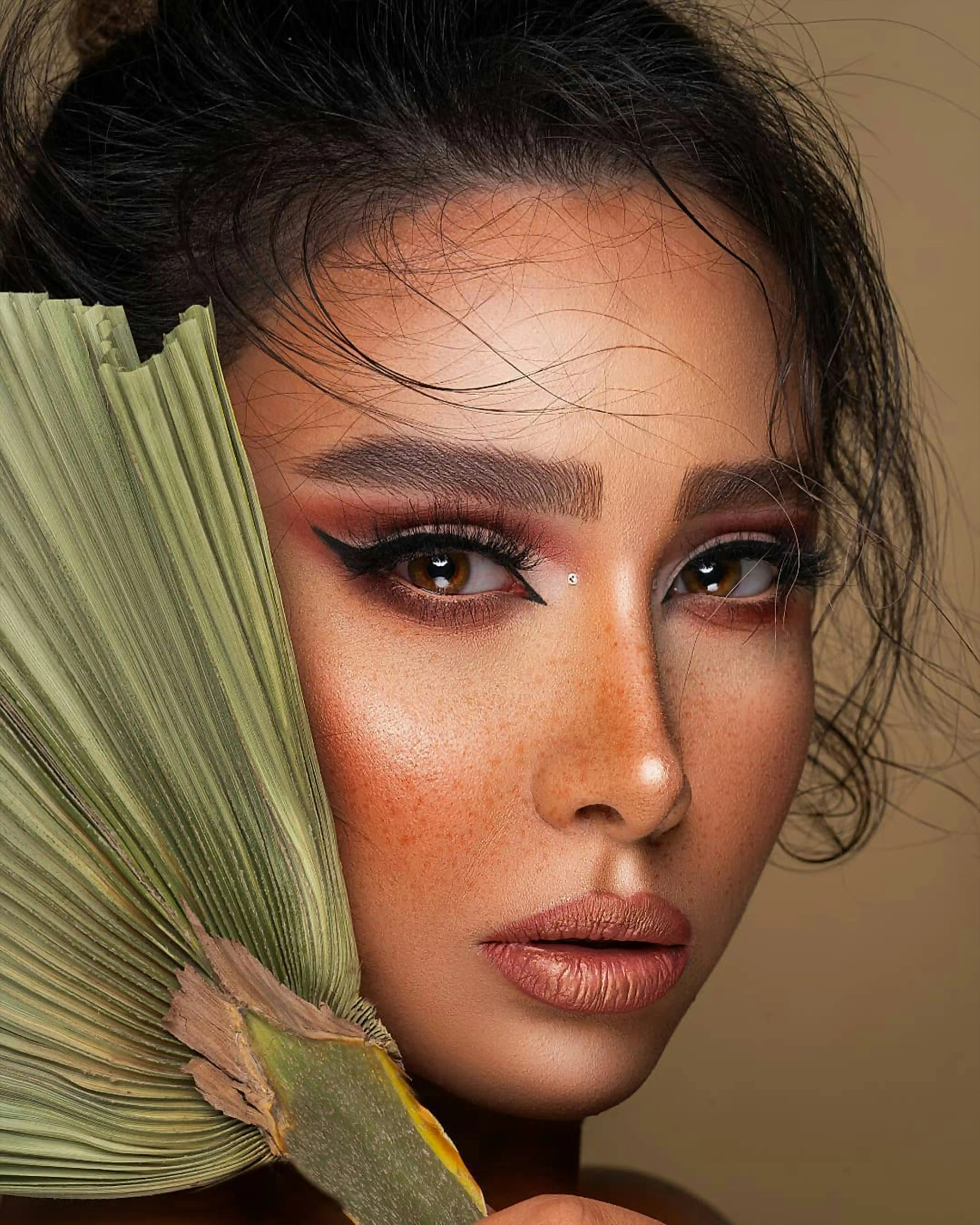 Cover Page model holding green fan near face, brown eyeshadow and freckles, thoughtful pose on neutral background.