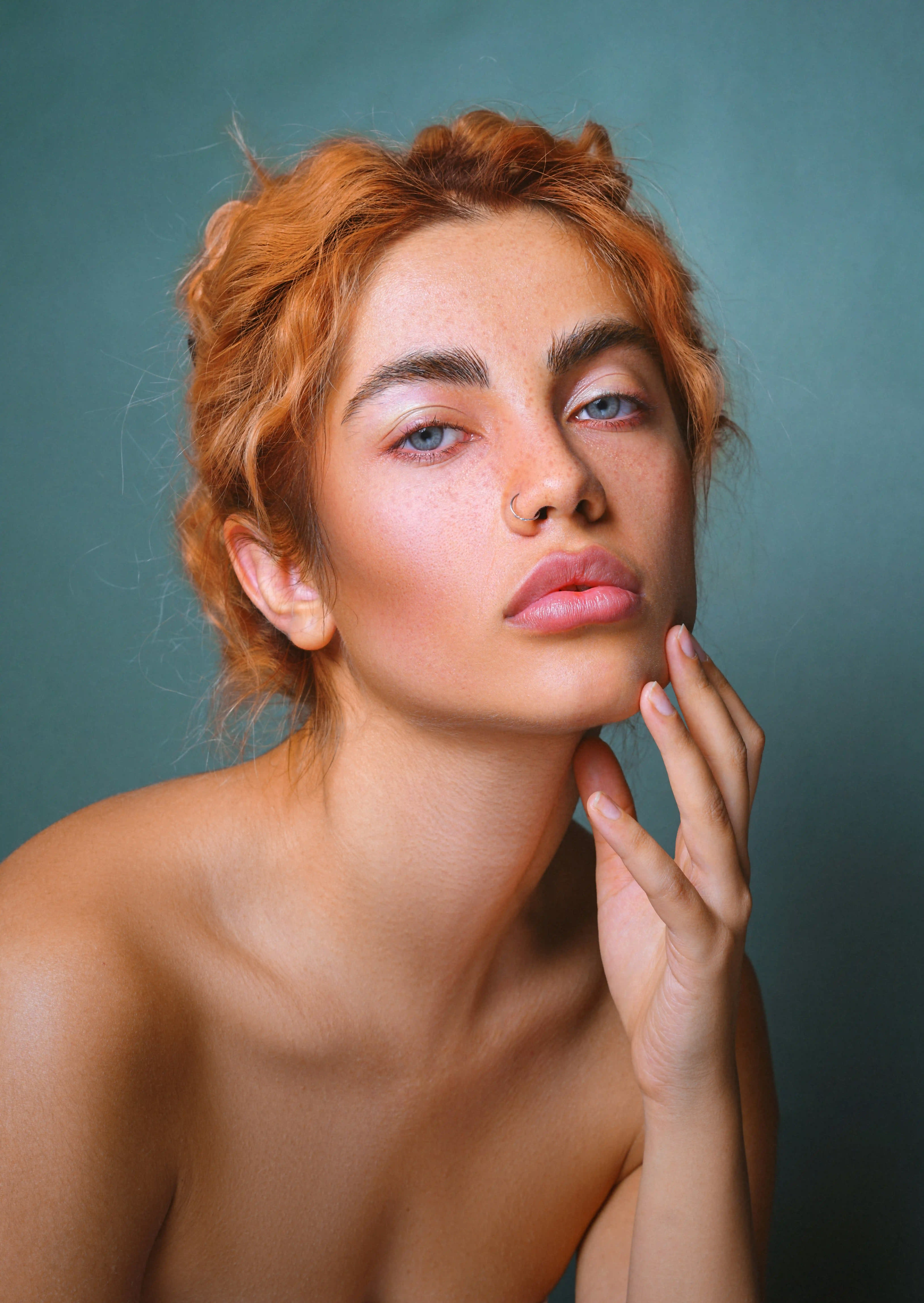 Cover Page model with curly red hair and nose ring, hand on chin with natural makeup, on teal background.
