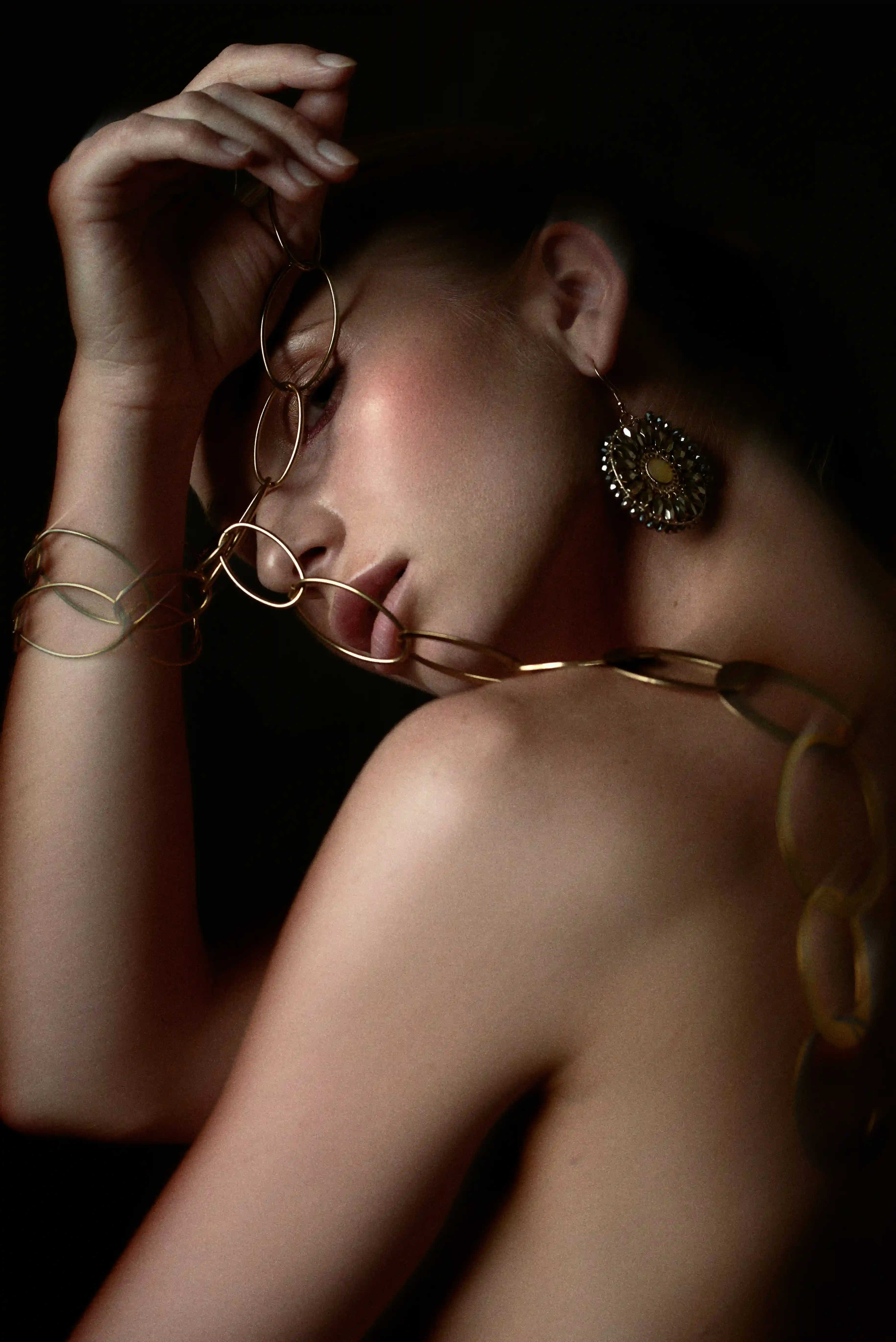 Cover Page model holding gold chain jewelry near face, with large earring and dramatic dark lighting, close-up portrait.