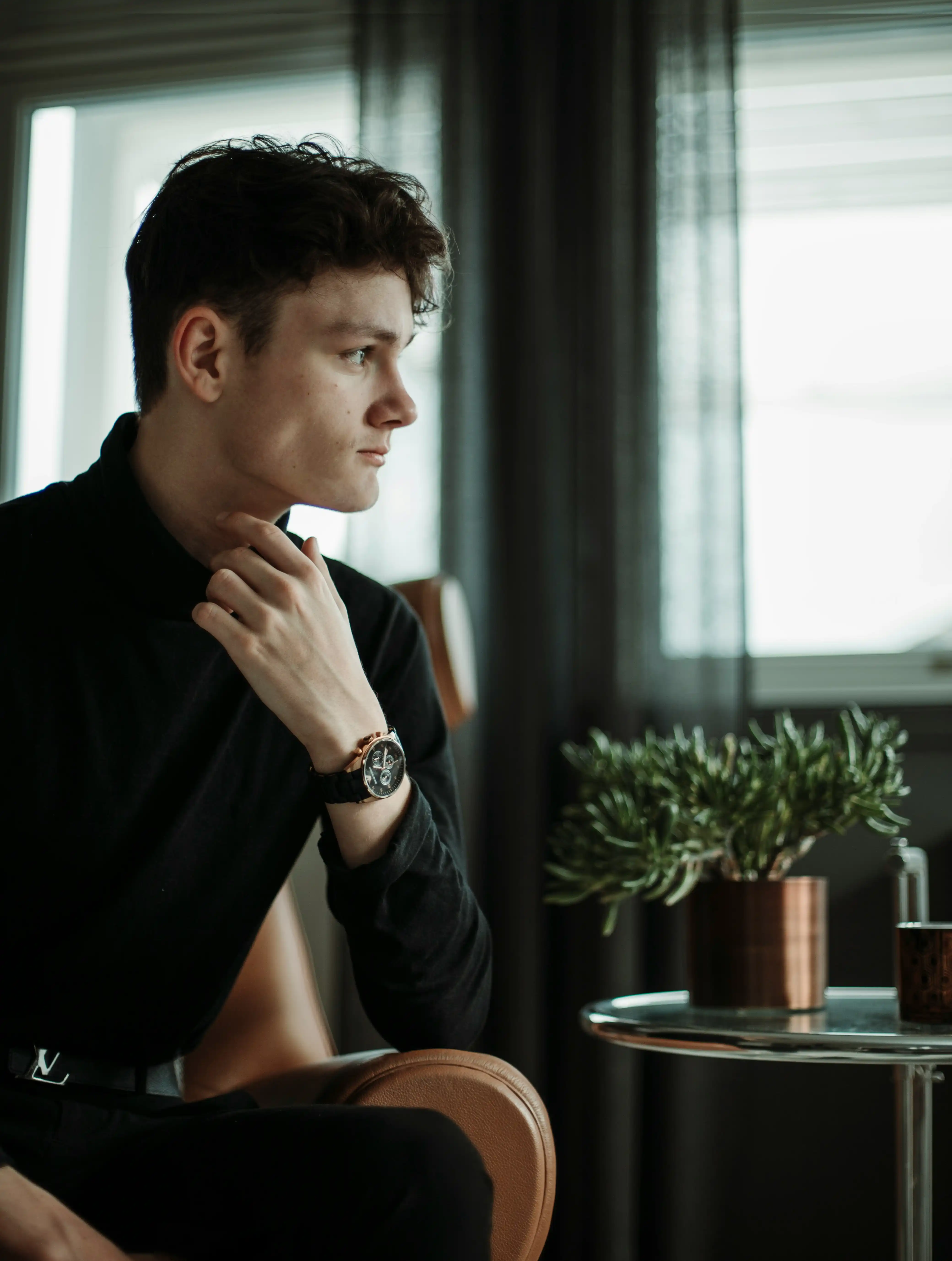 Cover Page thoughtful male model in black turtleneck, sitting in armchair by window, hand on chin with watch and indoor plants.