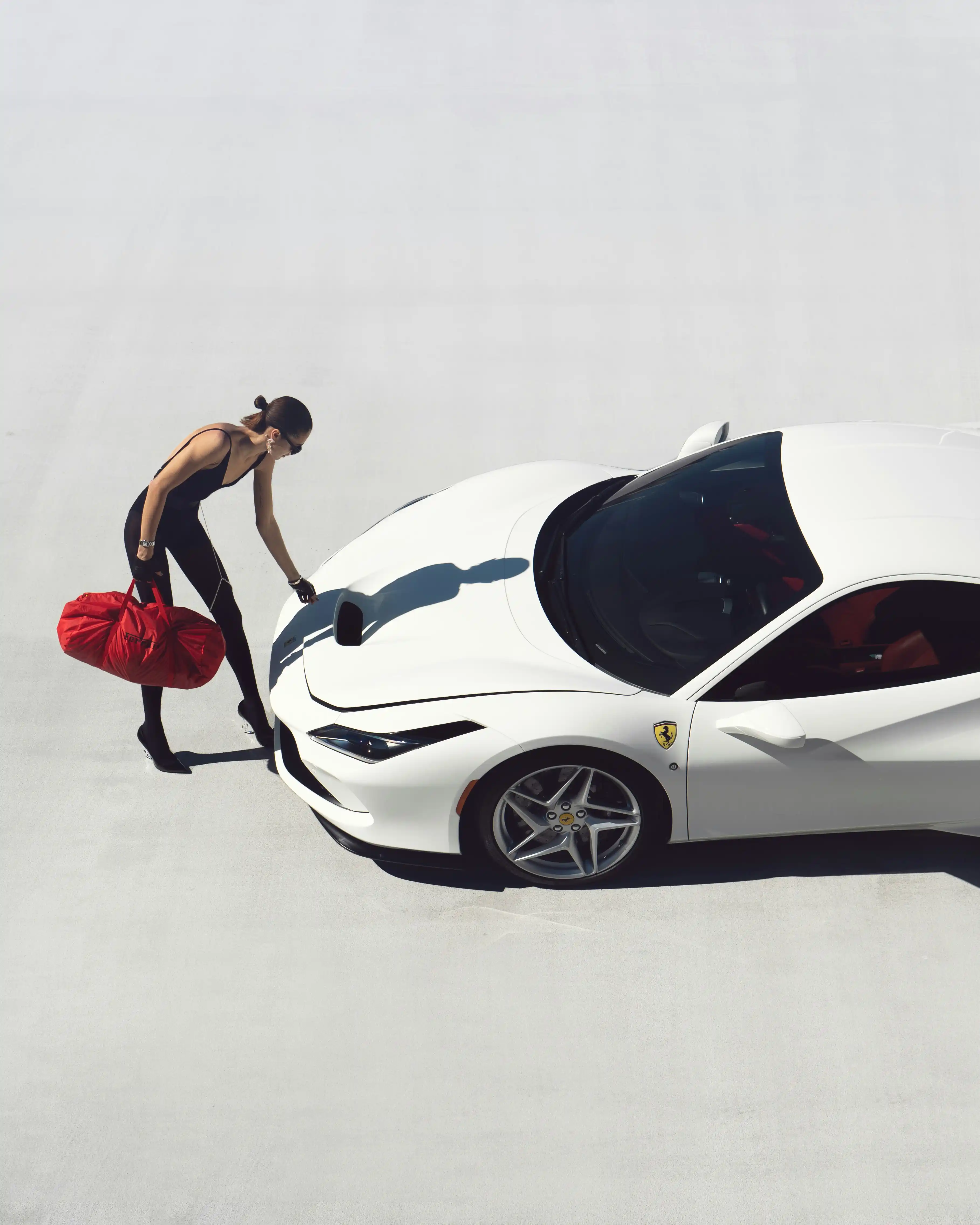 Cover Page model in black bodysuit and sunglasses, leaning on white Ferrari sports car with red tote bag in minimalist white setting.