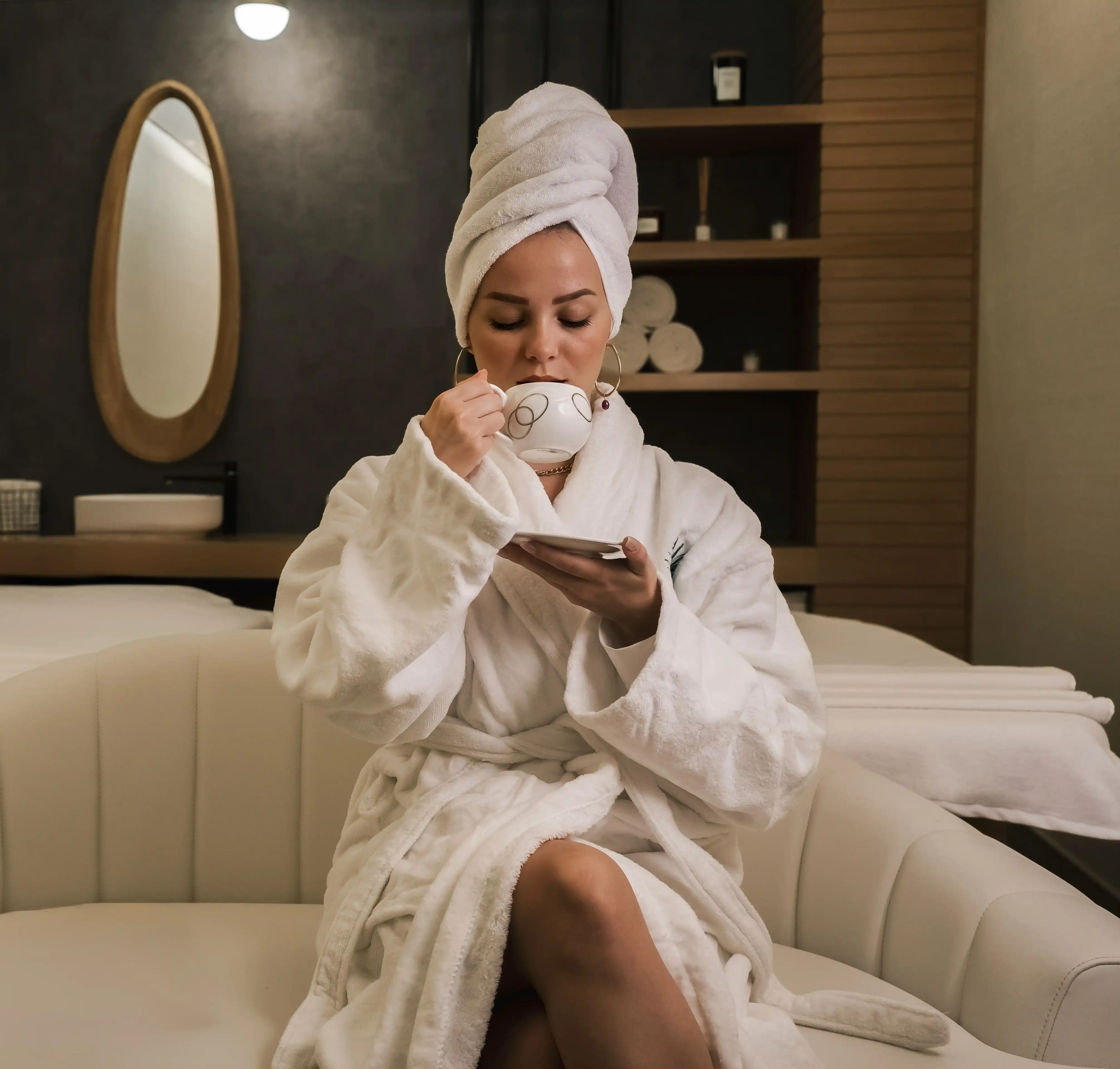 Cover Page relaxed model in white bathrobe and towel turban, sipping tea while checking phone on spa couch with mirror and shelves.