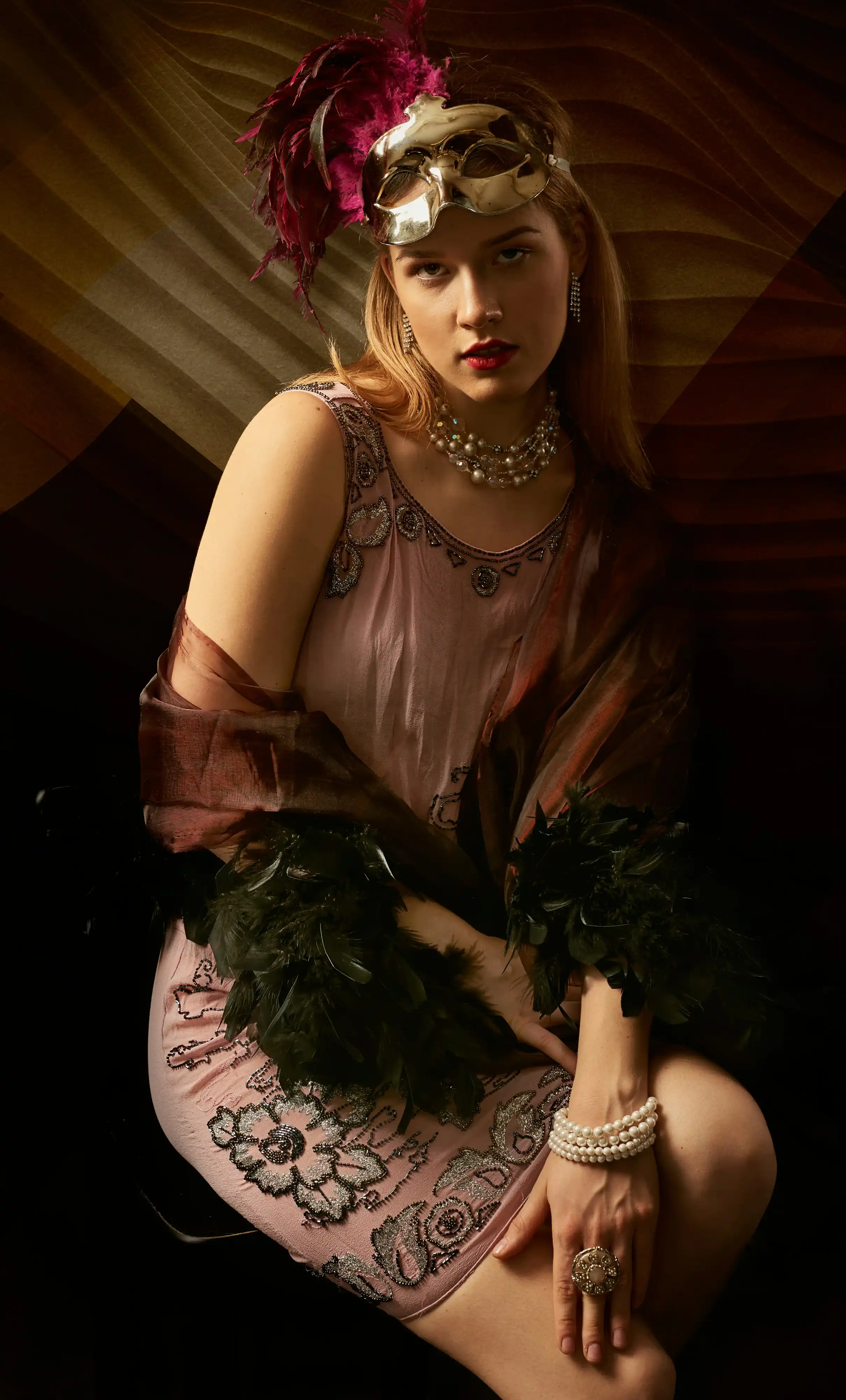 Cover Page model in pink beaded dress with feather boa and masquerade mask on head, pearl necklace and bracelet, seated in dramatic warm lighting.