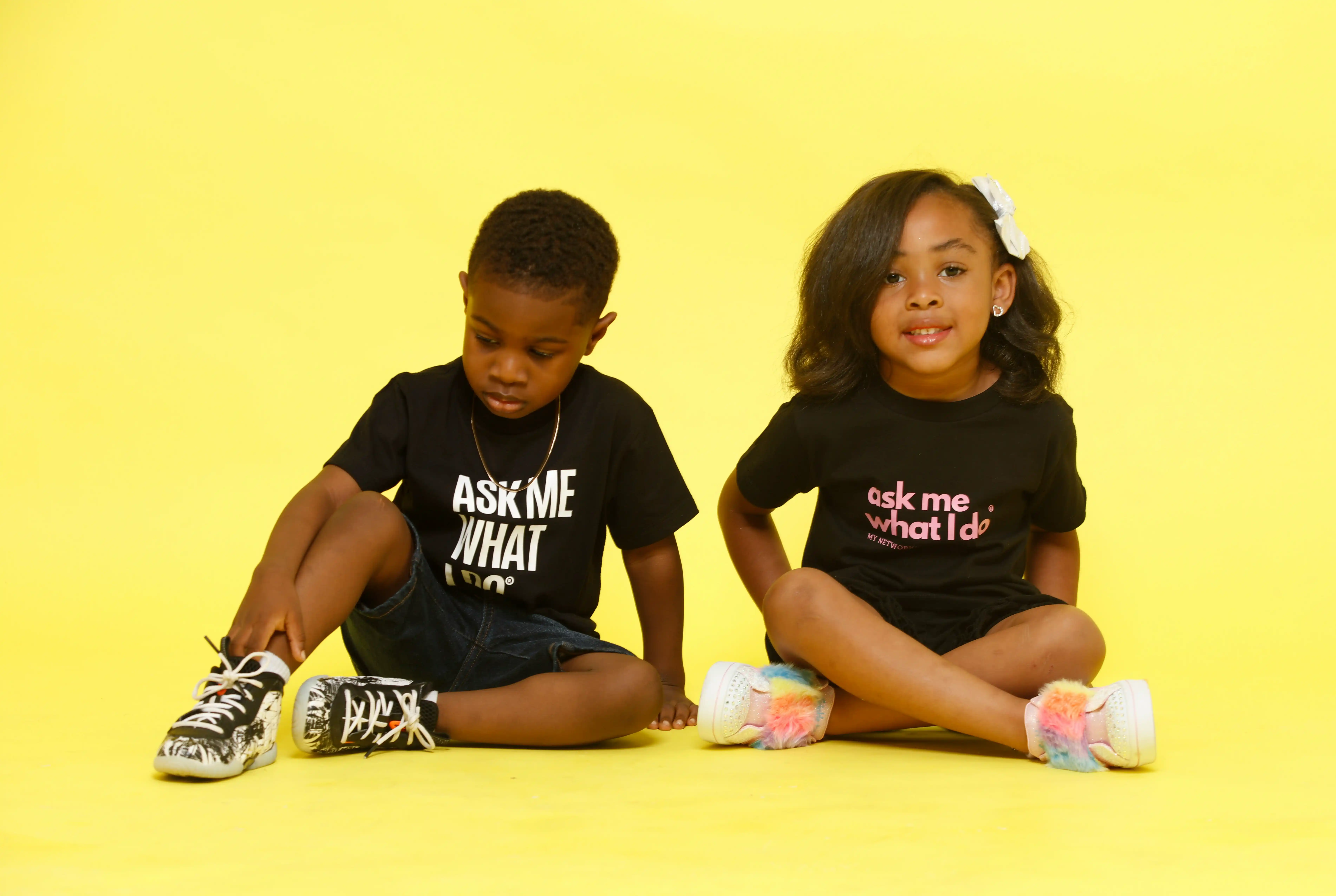 Cover Page two children sitting on yellow background, boy in black "ASK ME WHAT I DO" t-shirt tying sneaker, girl smiling in pink "ask me what I'd do" t-shirt with colorful shoes.