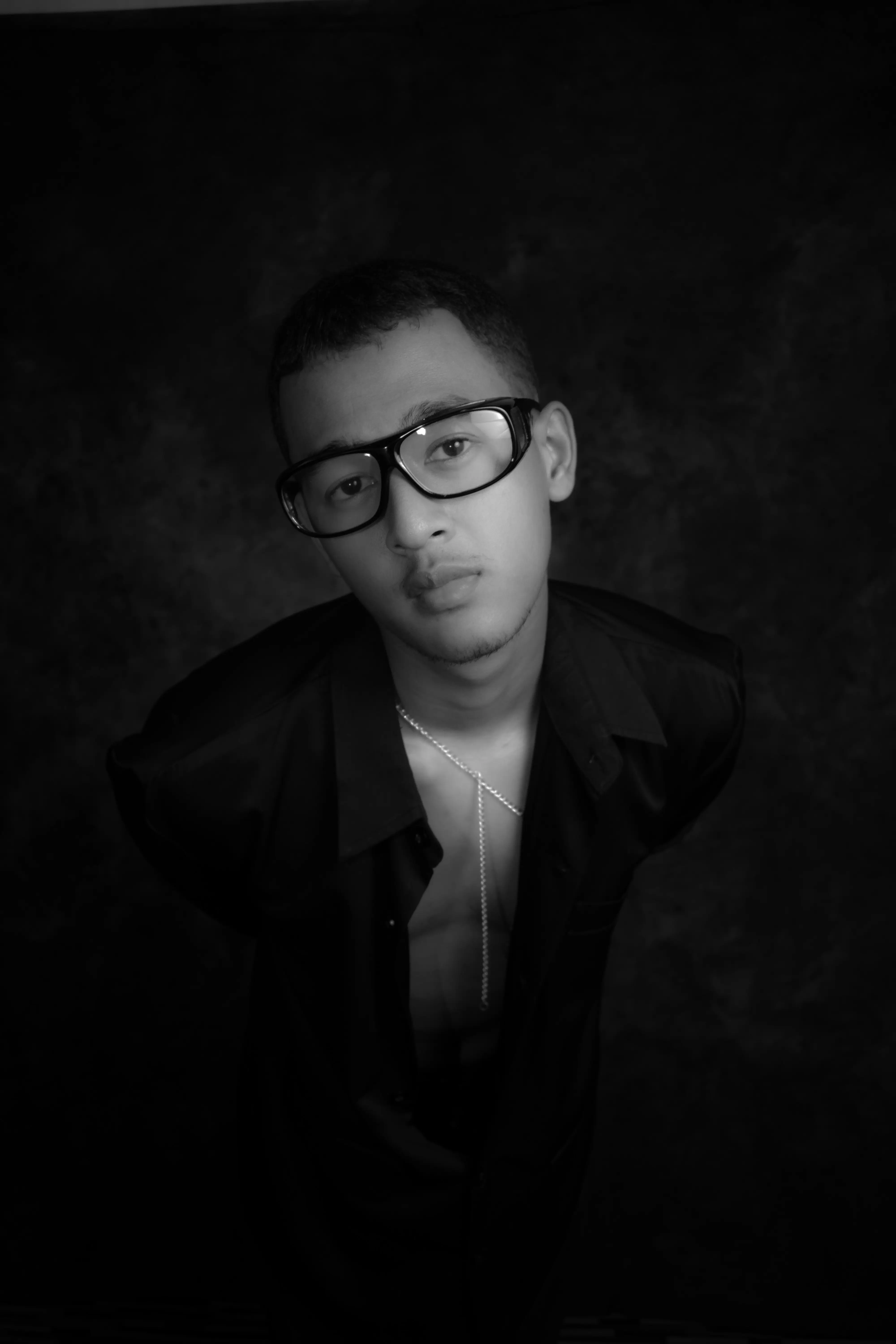 Cover Page young male model in black shirt and glasses, serious upward gaze with silver necklace, black and white portrait on dark background.