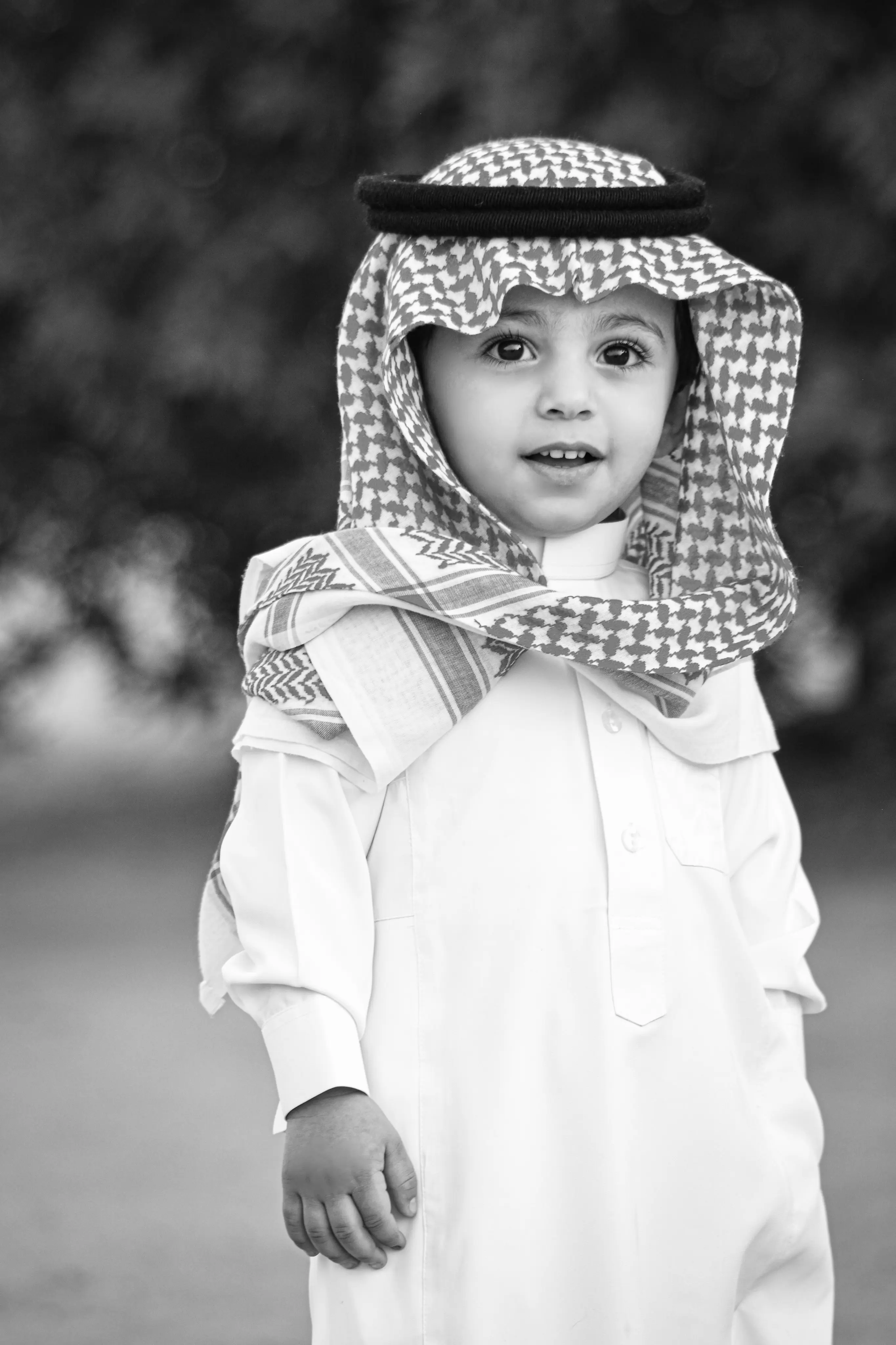 Cover Page young boy in white thobe and keffiyeh headscarf, smiling with hand down, black and white outdoor portrait on blurred background.