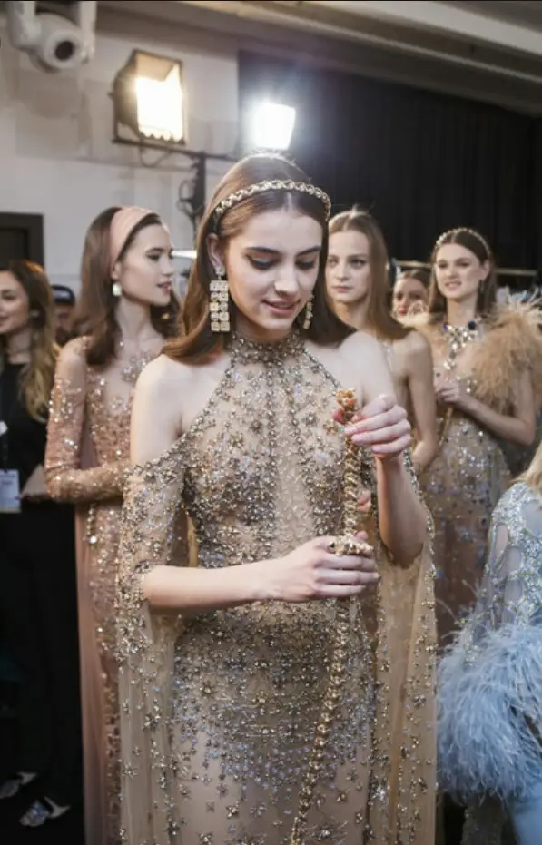 Group of female models in sparkling gold dresses backstage at a fashion show, one model in the foreground adjusting a necklace with an ornate headpiece.