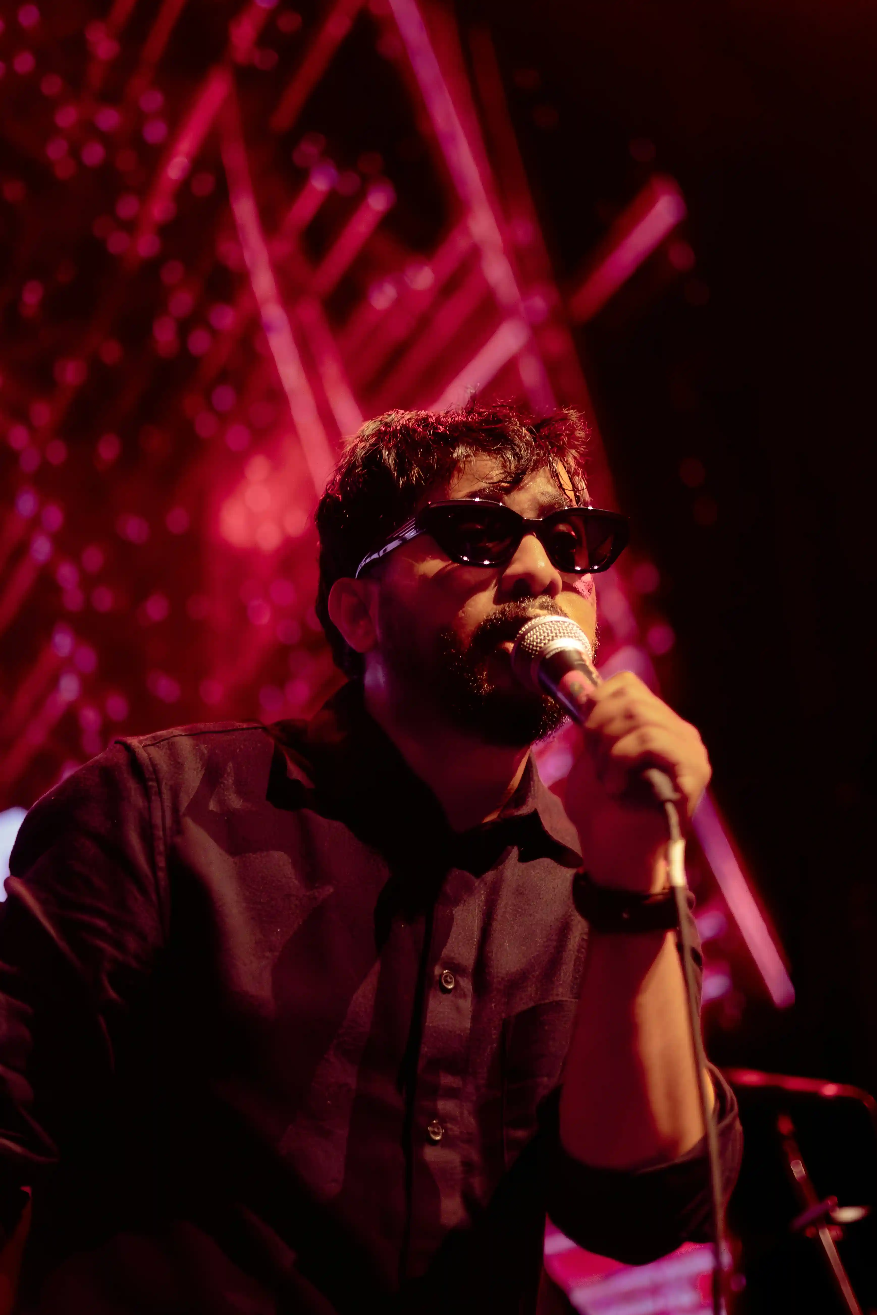 Male singer performing energetically on stage with microphone, wearing dark sunglasses and black shirt, dramatic red and pink stage lighting with illuminated tower backdrop during live entertainment event in Dubai