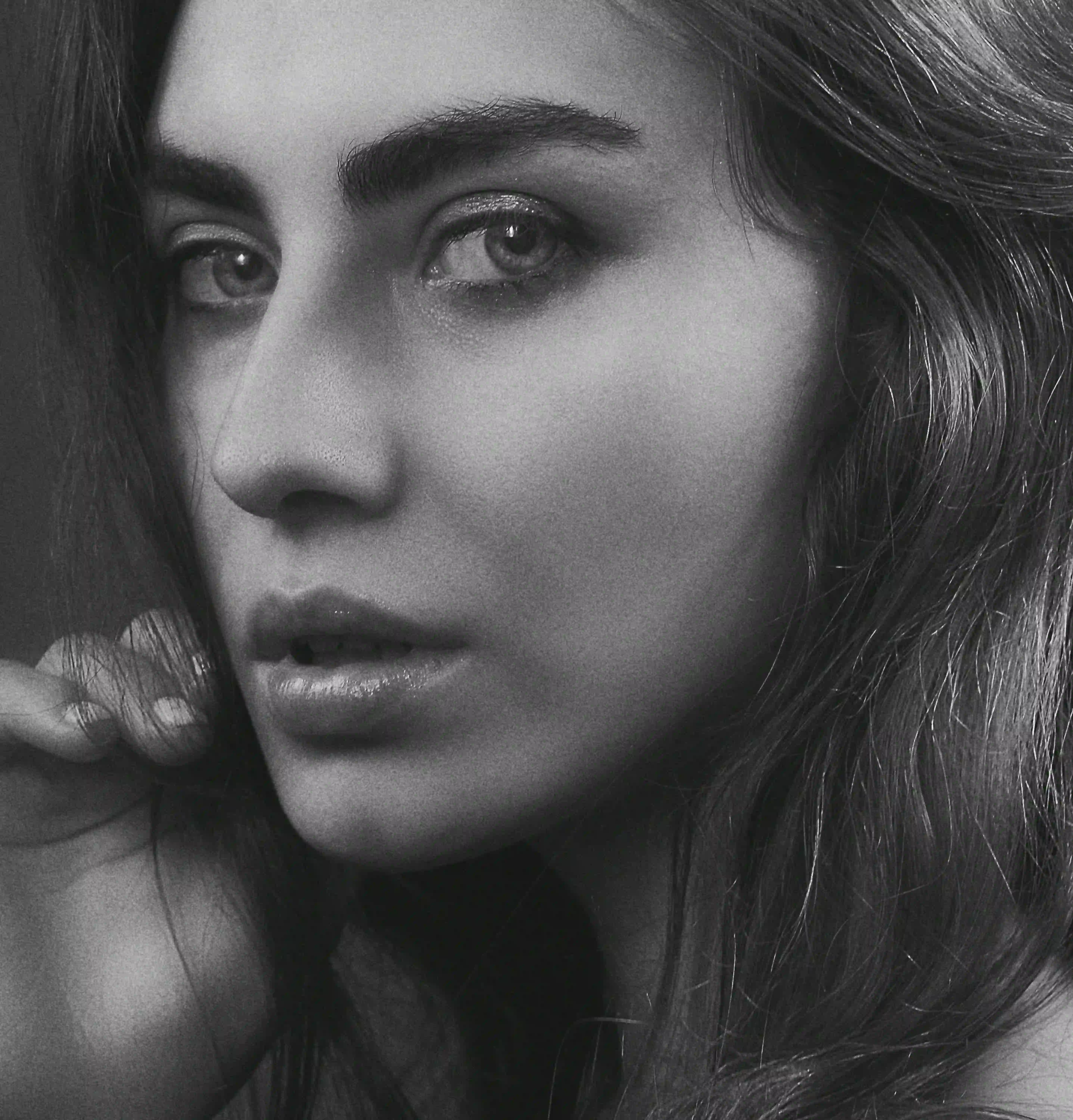 Intense black and white close-up portrait of woman with long dark hair, hand touching strands near face, glossy lips and striking eyes in dramatic low-key lighting, seductive high-fashion beauty shot