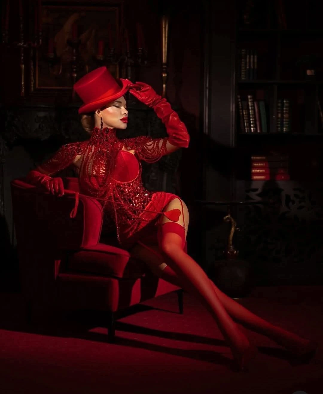 Glamorous woman in sparkling red burlesque cabaret outfit with top hat and long gloves, sensual pose on velvet armchair in luxurious library setting, Dubai nightlife elegance and high-end entertainment 2026