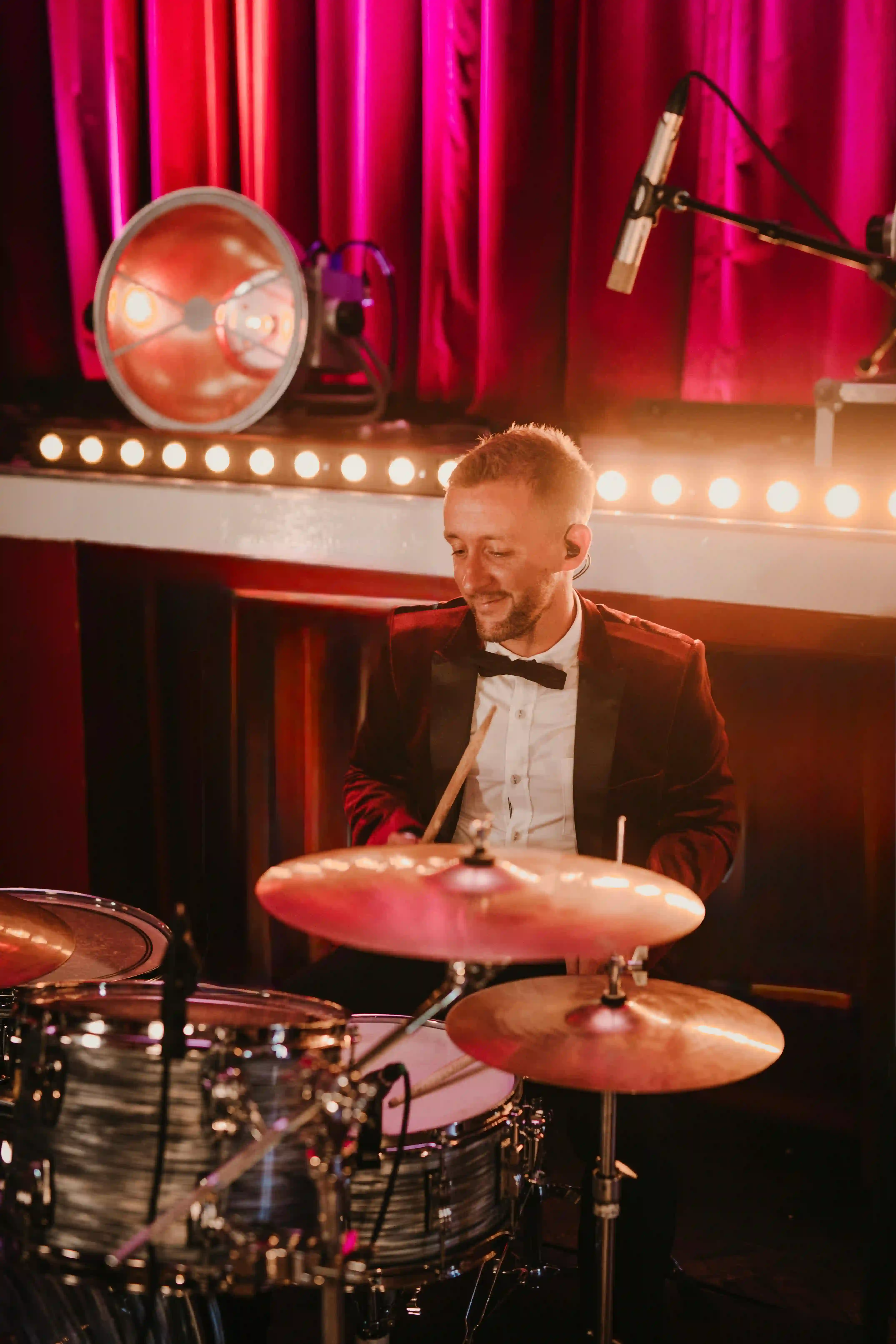 Professional drummer in elegant maroon velvet tuxedo and bow tie passionately playing drum kit on stage under vibrant pink and purple lights, live music entertainment for Dubai luxury events, weddings, corporate galas, jazz nights and dinner shows 2026