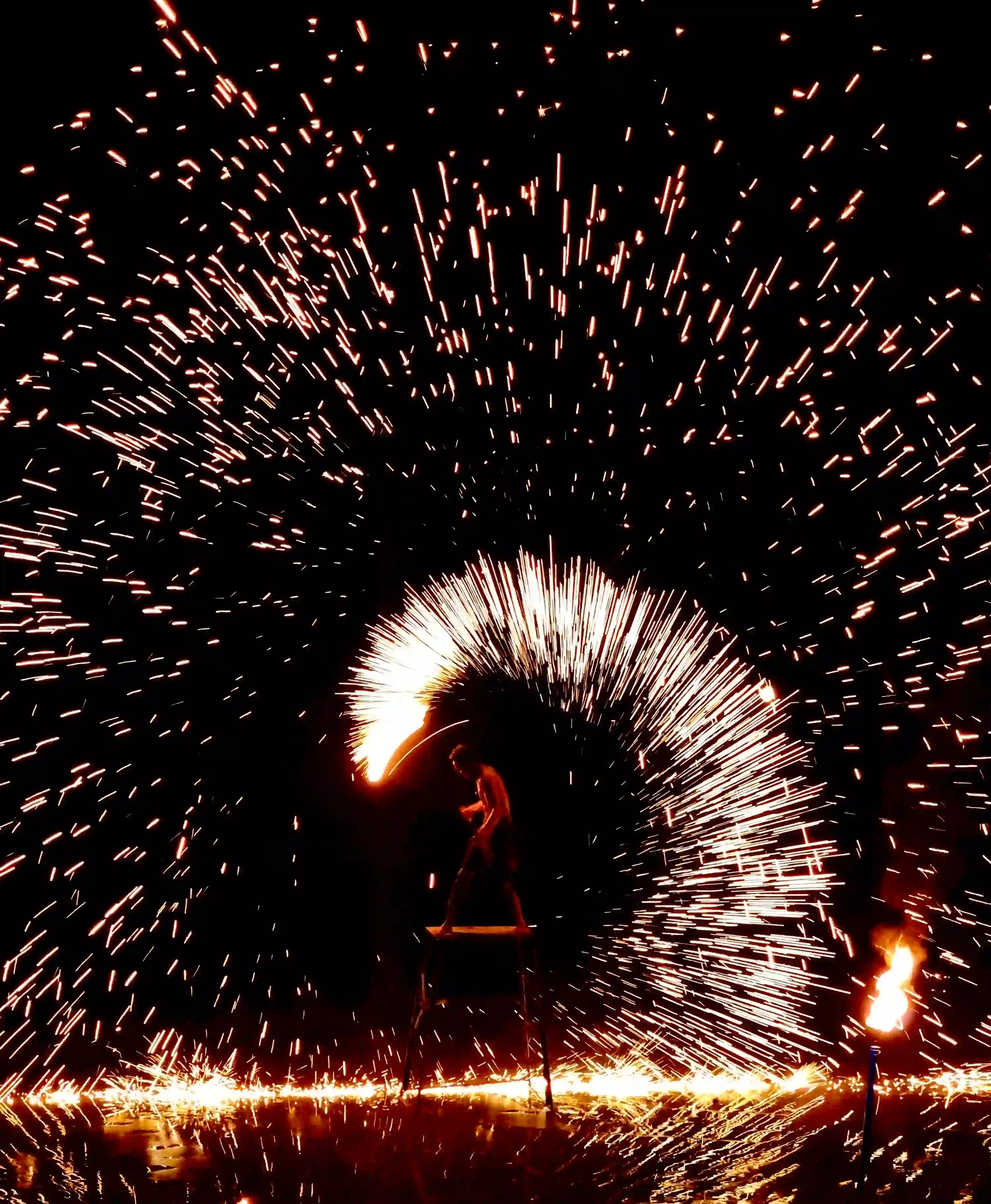 Dramatic fire performer on ladder creating huge peacock fan of bright orange sparks and flames at night, intense sparks flying in dark sky, spectacular live fire show entertainment for Dubai luxury events, weddings, corporate galas and desert parties 2026