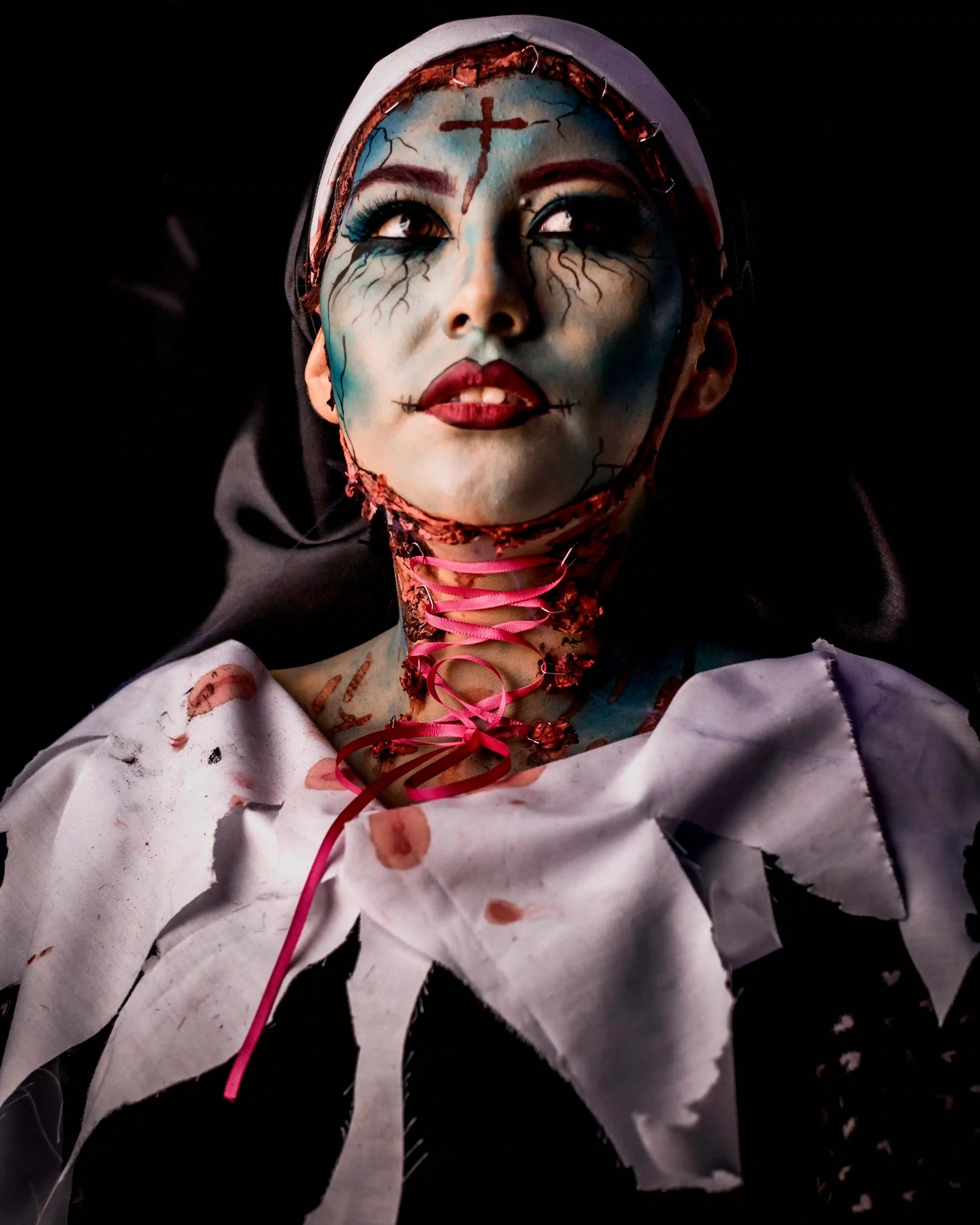 Terrifying evil nun in possessed horror makeup: blue cracked skin, stitched forehead cross, black veins, red lips, and pink ribbon-laced bloody neck. Dramatic close-up performance by Cover Page Agency Dubai. Ideal for Halloween events, themed parties, luxury nightlife, and spooky activations in UAE.