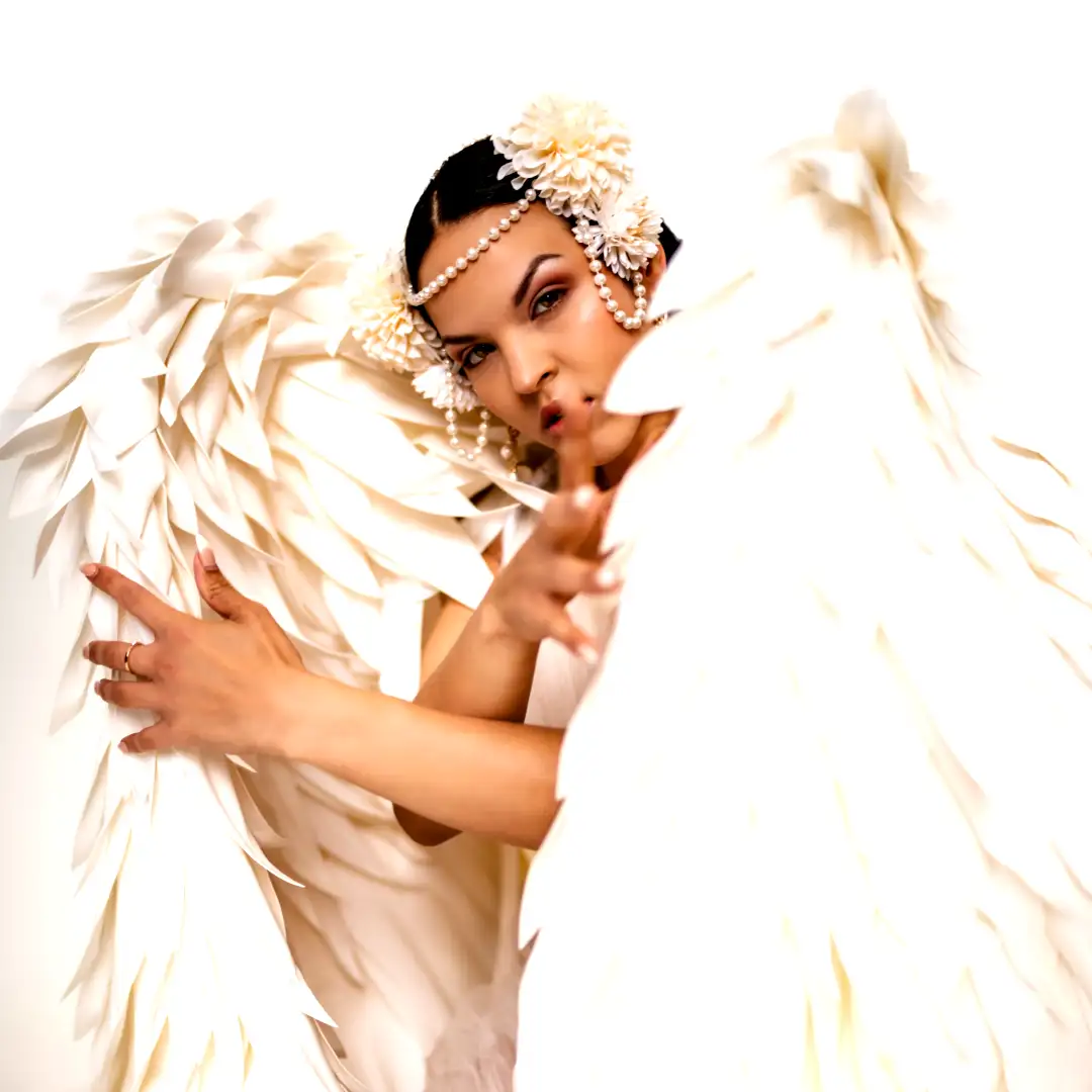 Woman in luxurious white angel wings costume with large folded fabric wings, pearl and floral headpiece, dramatic close-up pose for entertainment photoshoot