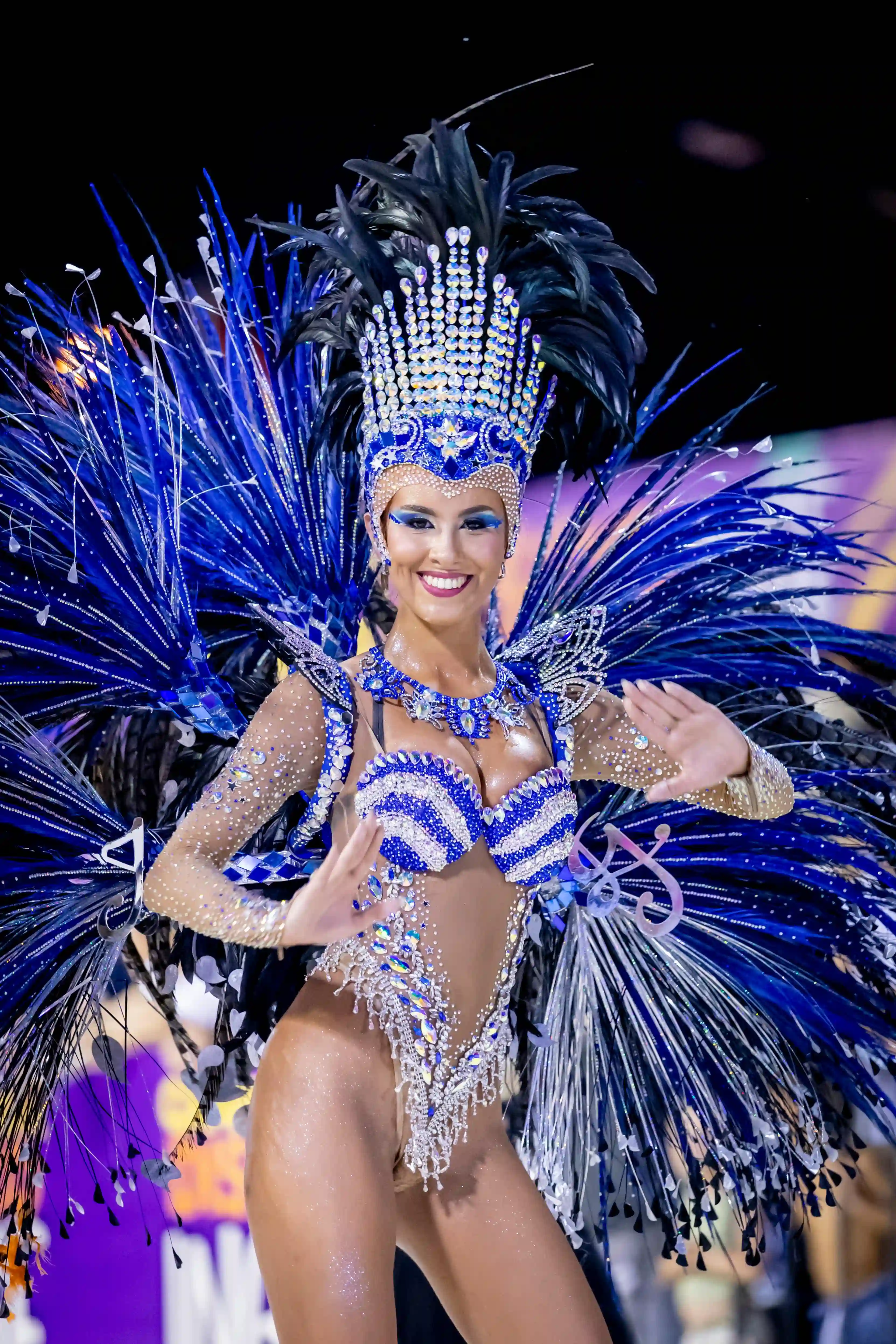 Stunning luxury samba dancer in an elaborate blue and black feathered carnival costume with sparkling rhinestones smiling confidently for Cover Page Agency Dubai. High-end Brazilian carnival entertainment talent delivering vibrant, glamorous and high-energy performances at exclusive events and luxury parties across the UAE.