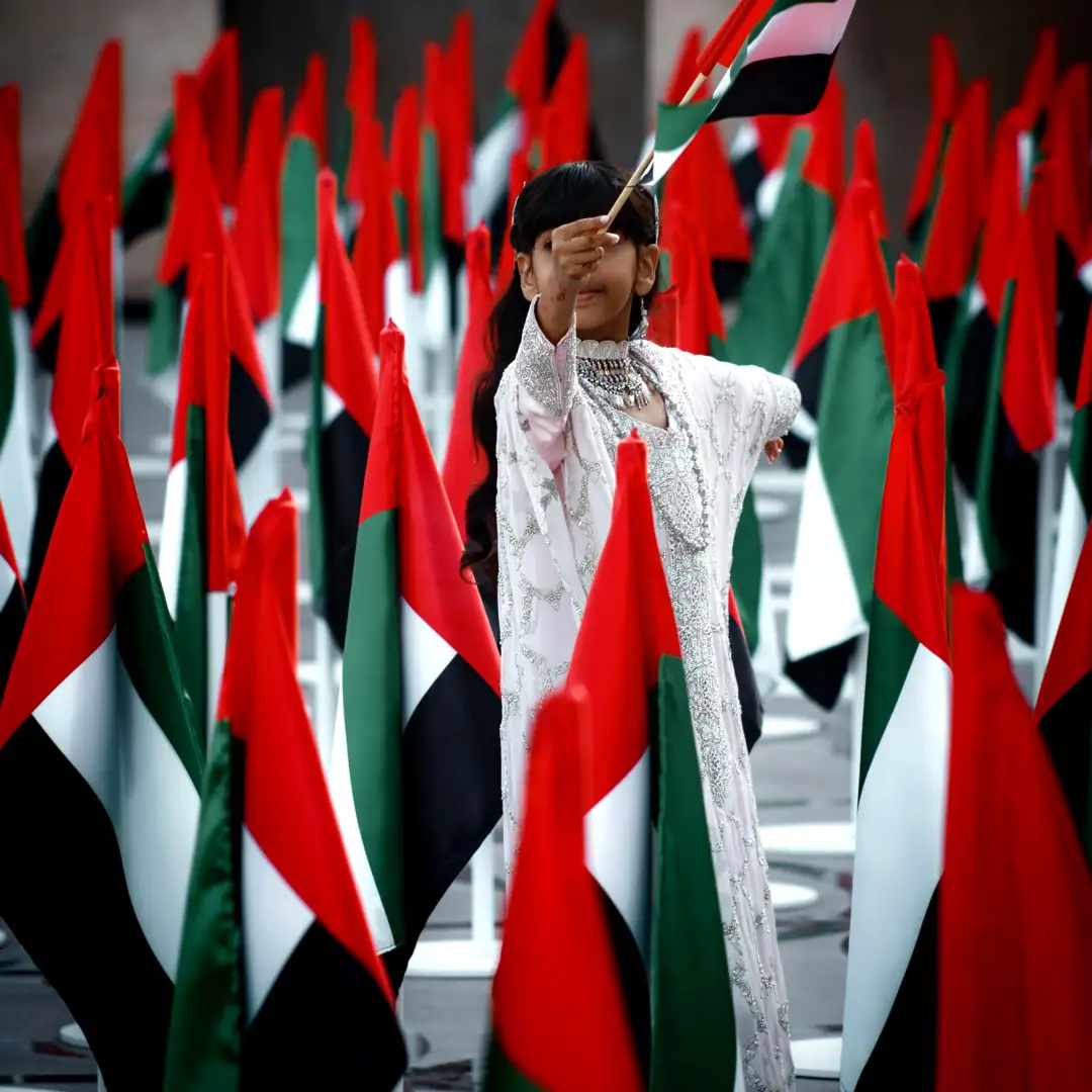 Elegant young performer in traditional white Emirati attire proudly waving the UAE flag among a sea of national flags for Cover Page Agency Dubai. High-end cultural entertainment talent delivering patriotic, graceful and visually stunning performances at exclusive national day events, luxury galas and celebrations across the UAE.
