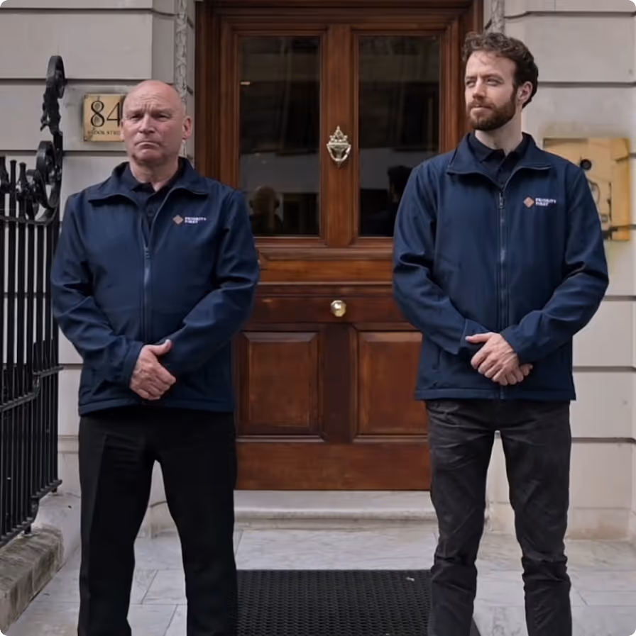 Two men in navy jackets standing in front of a wooden door with the number 84 on the wall.