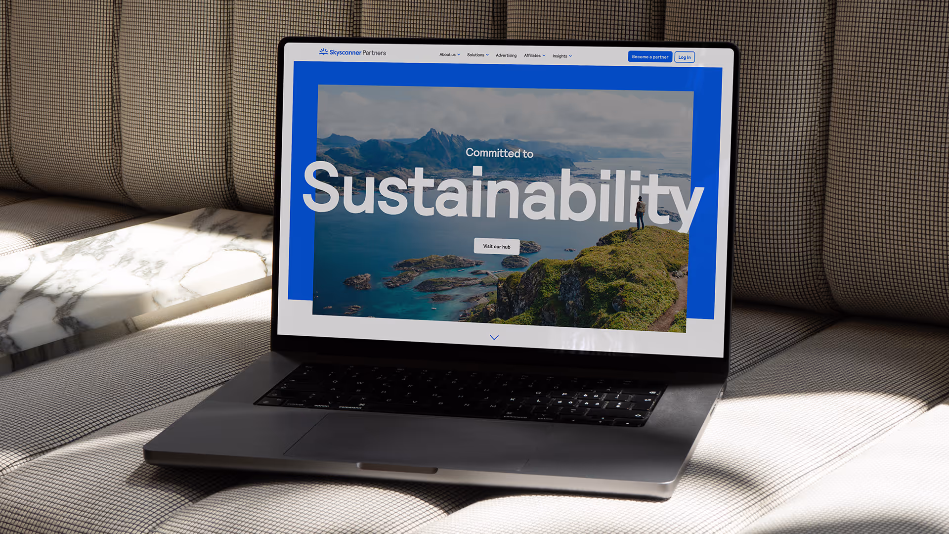 Laptop on checkered couch showing a webpage with 'Committed to Sustainability' over a coastal landscape.