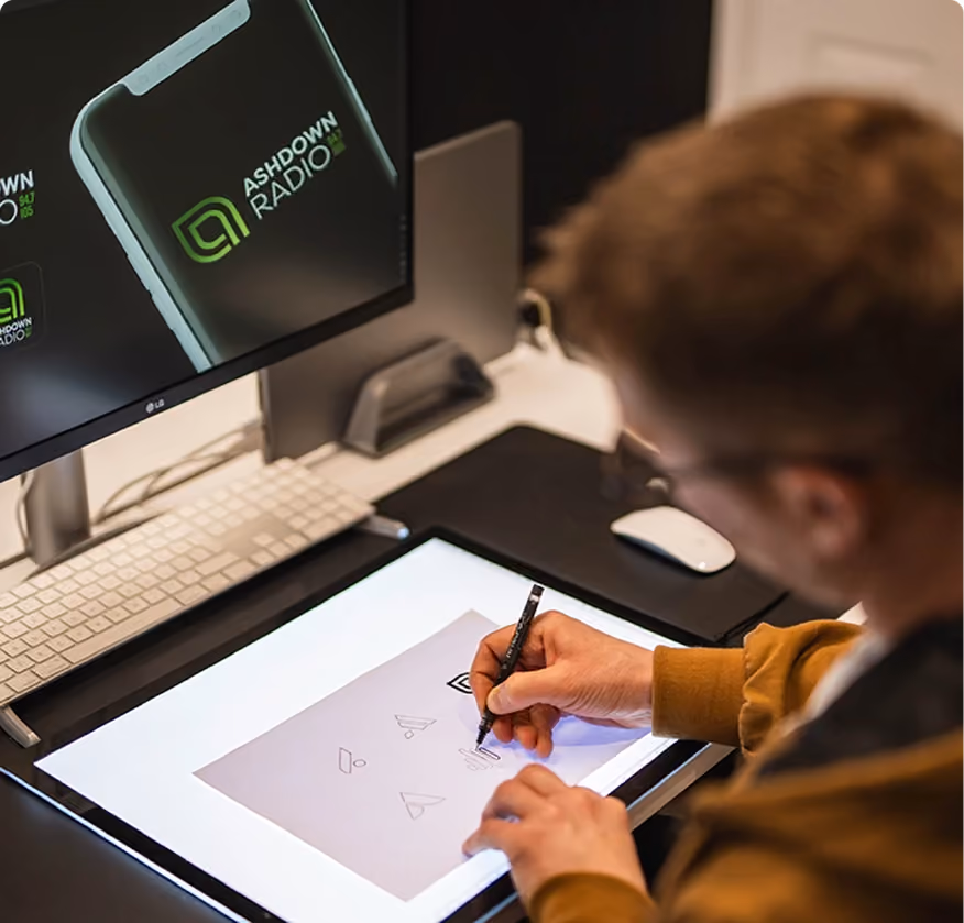 Person sketching logo designs on a lightbox with a computer screen showing Ashdown Radio logo nearby.