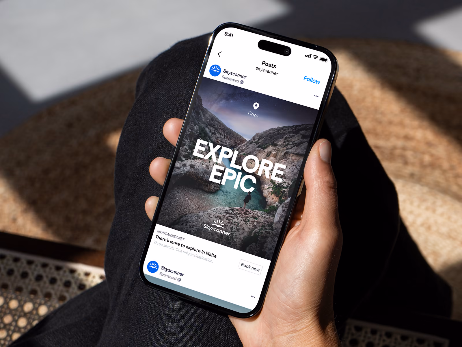 Hand holding smartphone displaying Skyscanner ad with 'Explore Epic' over rocky coastal scenery.