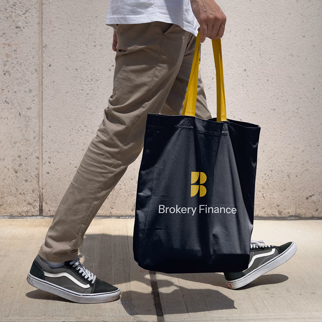 Person walking wearing black and white sneakers, carrying a black tote bag with yellow handles and Brokery Finance logo.