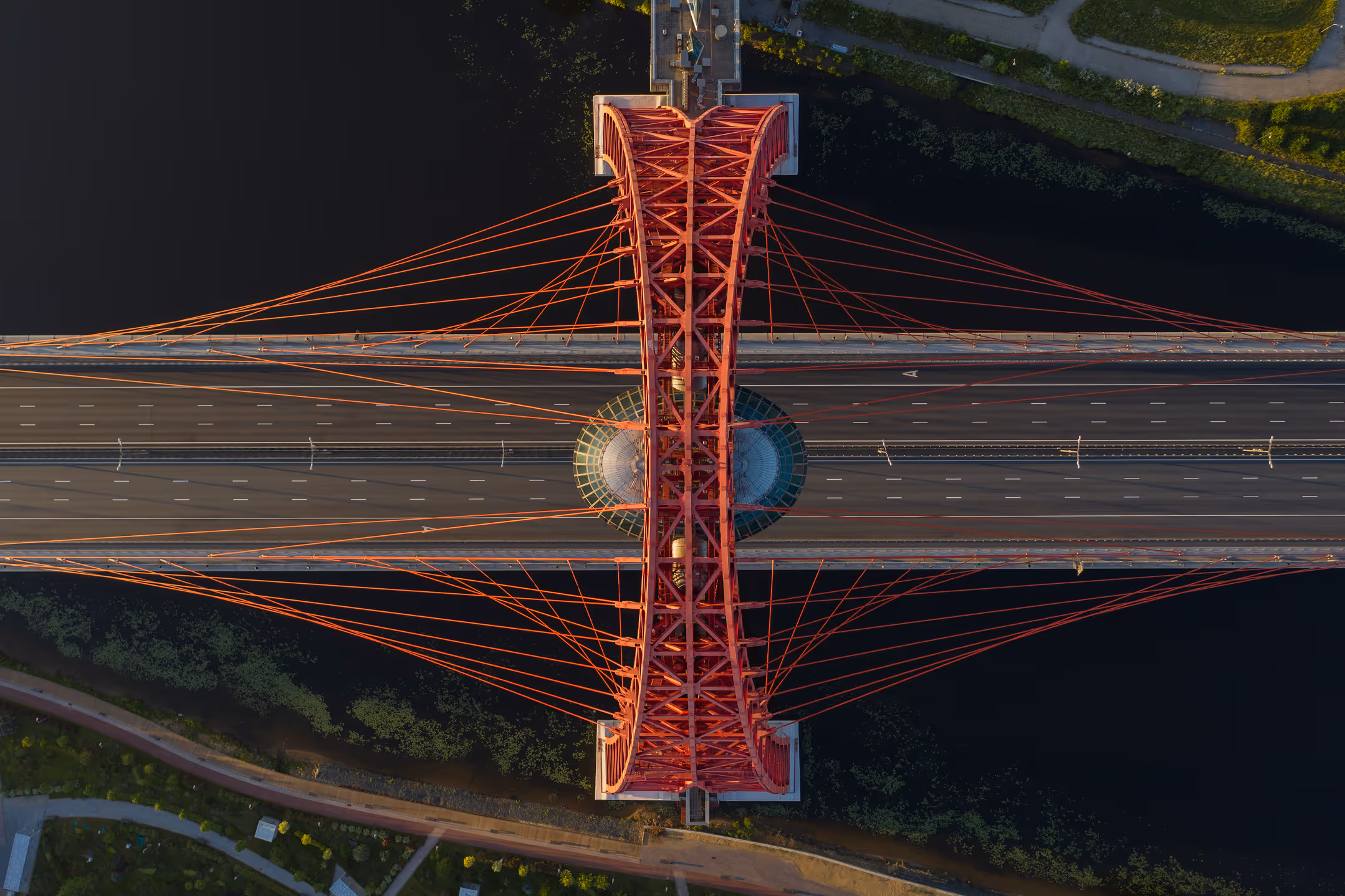 Aerial top-down view of a red cable-stayed bridge spanning over a dark river with multiple lanes and minimal traffic.