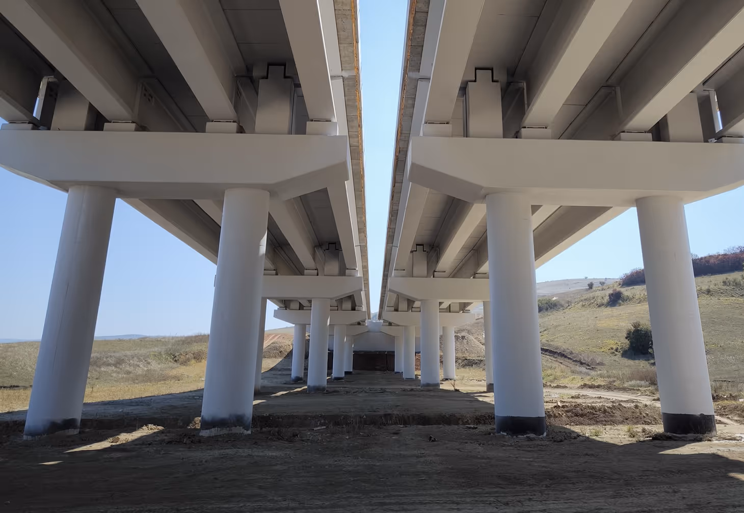 View underneath a concrete bridge with white cylindrical columns supporting two parallel roadway sections.