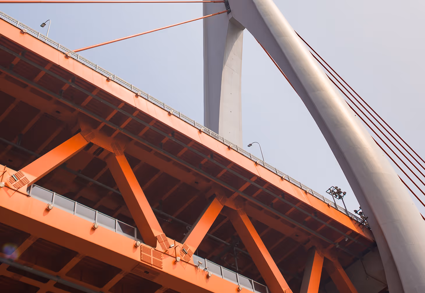 Close-up view of an orange metal bridge structure with large white cylindrical support beams and cables against a pale sky.