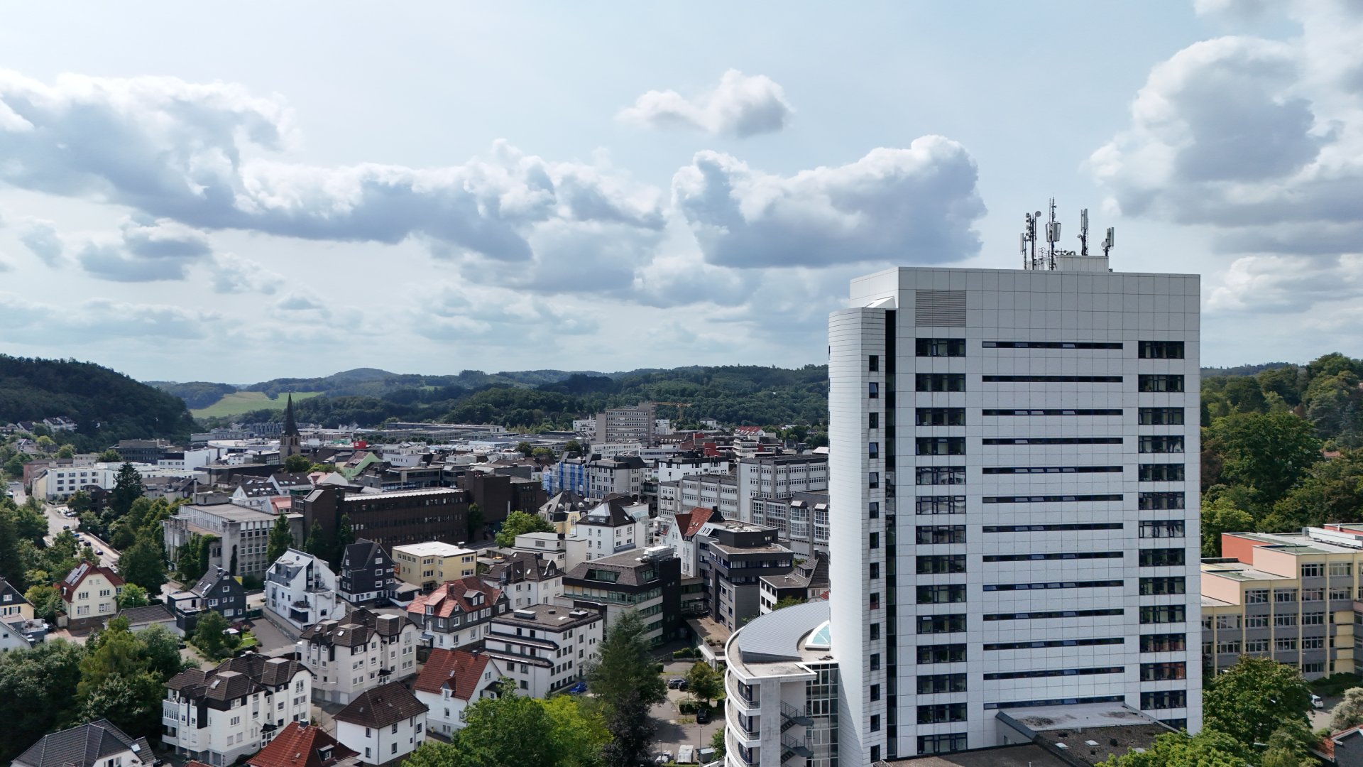 Cityscape featuring a tall modern white building with antennas, surrounded by smaller residential and commercial buildings with hills in the background under a partly cloudy sky.