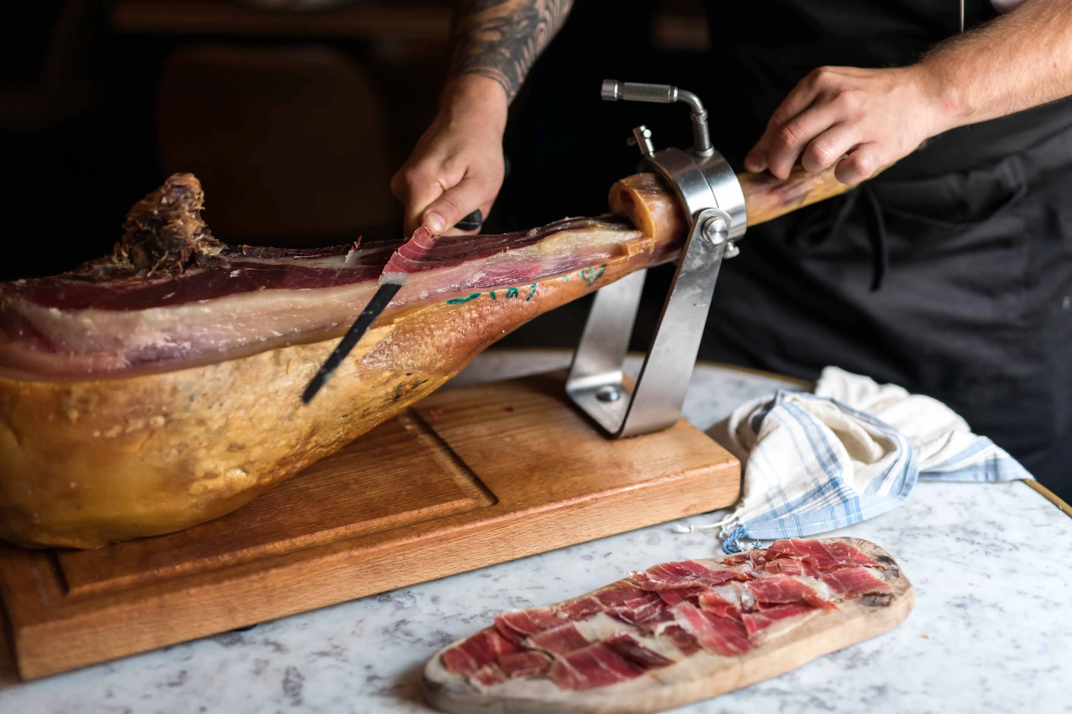 Person slicing thin pieces of cured ham from a leg mounted on a wooden stand with sliced ham arranged on a wooden board nearby.
