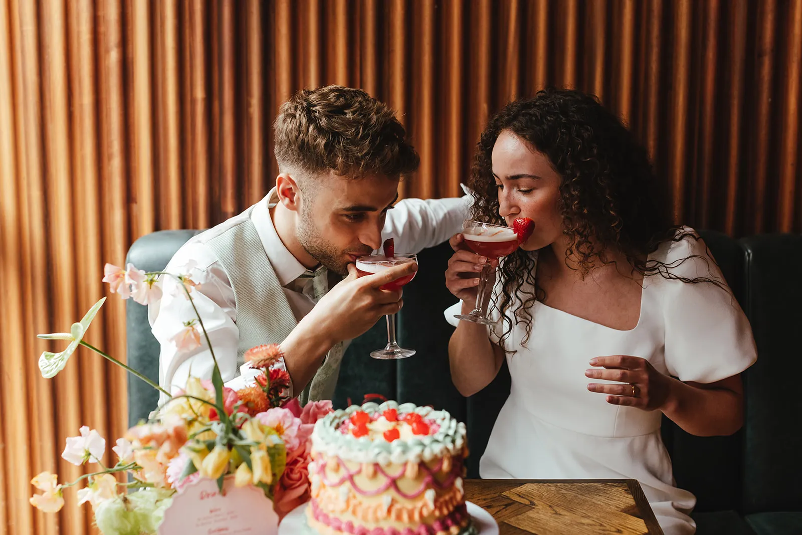 Couple dressed in wedding attire sipping red cocktails with strawberries, seated at a table with a decorated cake and floral arrangement.